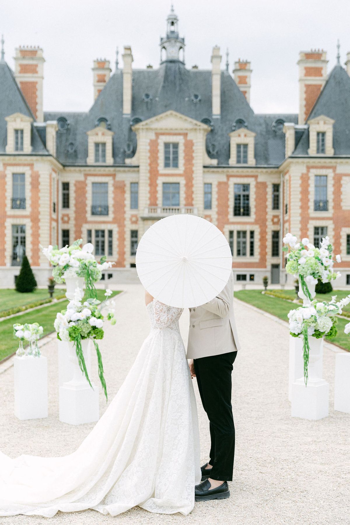 Couple under a white parasol on the gravel aisle between the ceremony's floral plinths