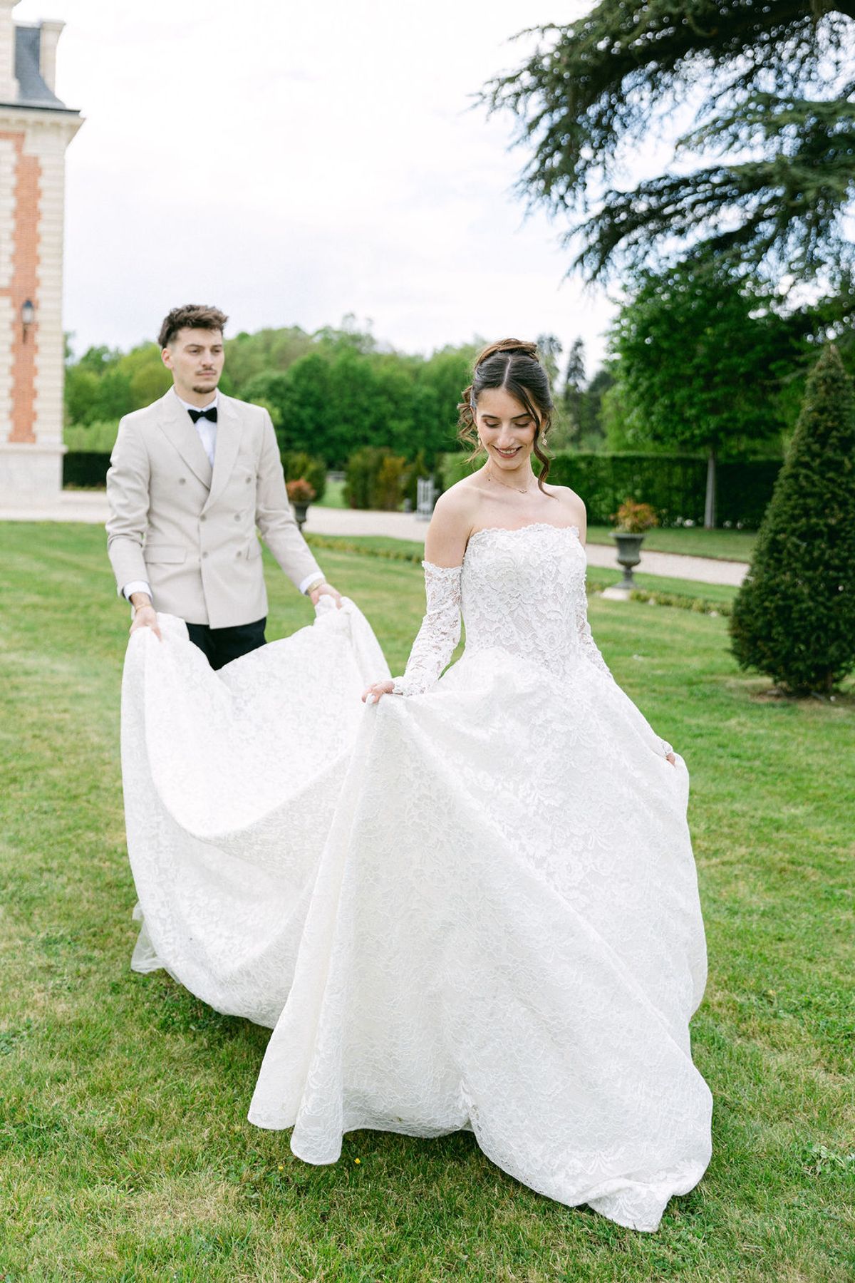 Groom carries the bride's train as they cross the lawn together