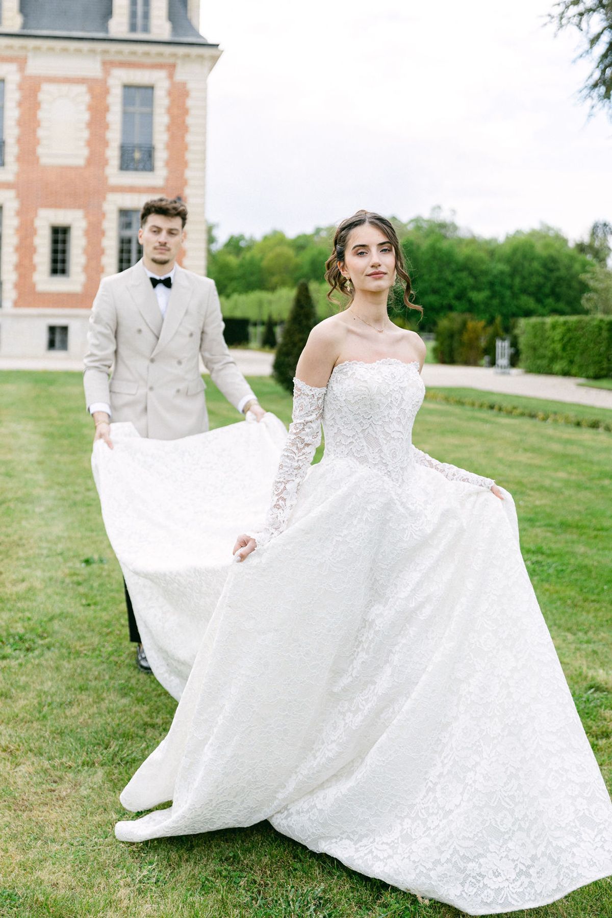 Groom lifts the bride's train as they walk across the lawn