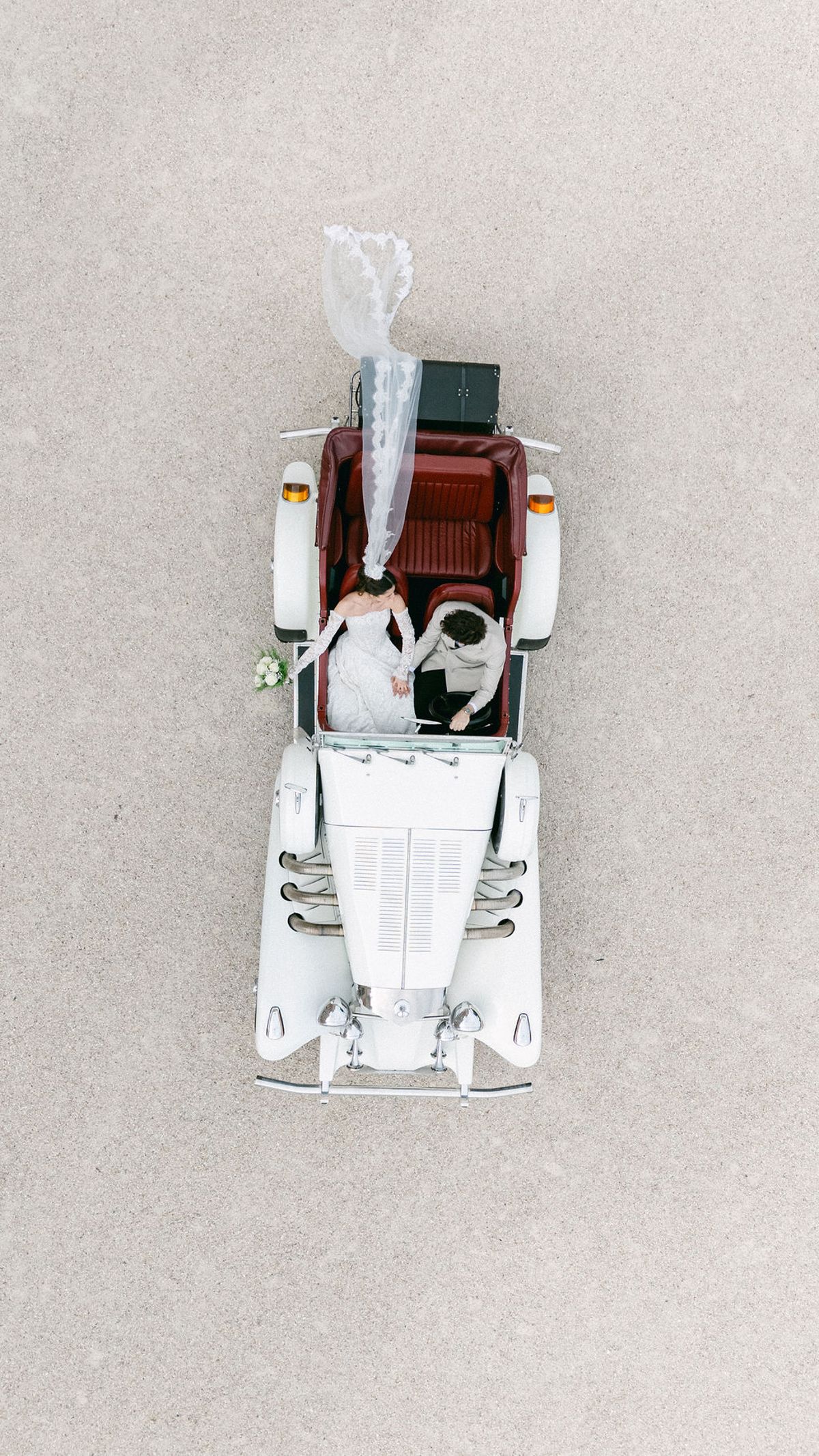 Aerial view straight down on the couple in the open vintage car, the bride's veil streaming up the gravel