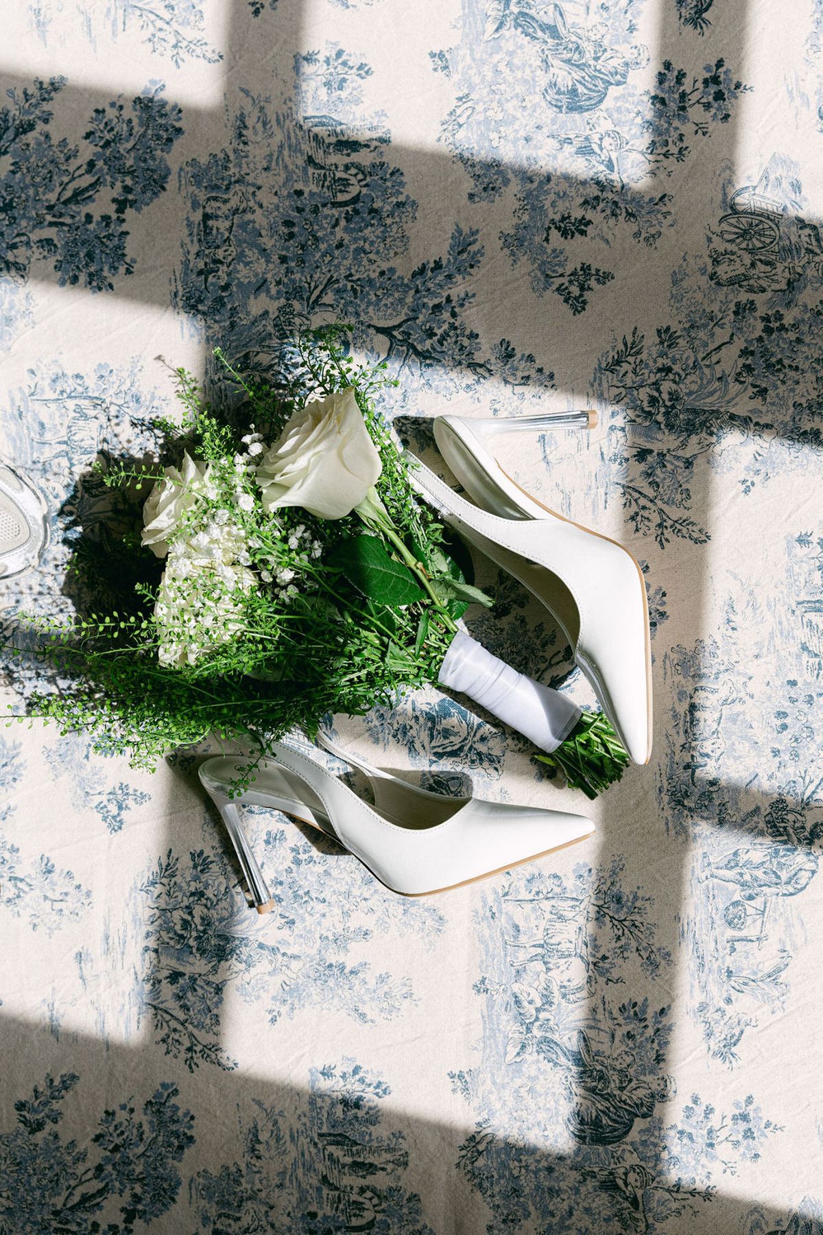 Bridal bouquet of white blooms and greenery beside white heels on blue toile fabric