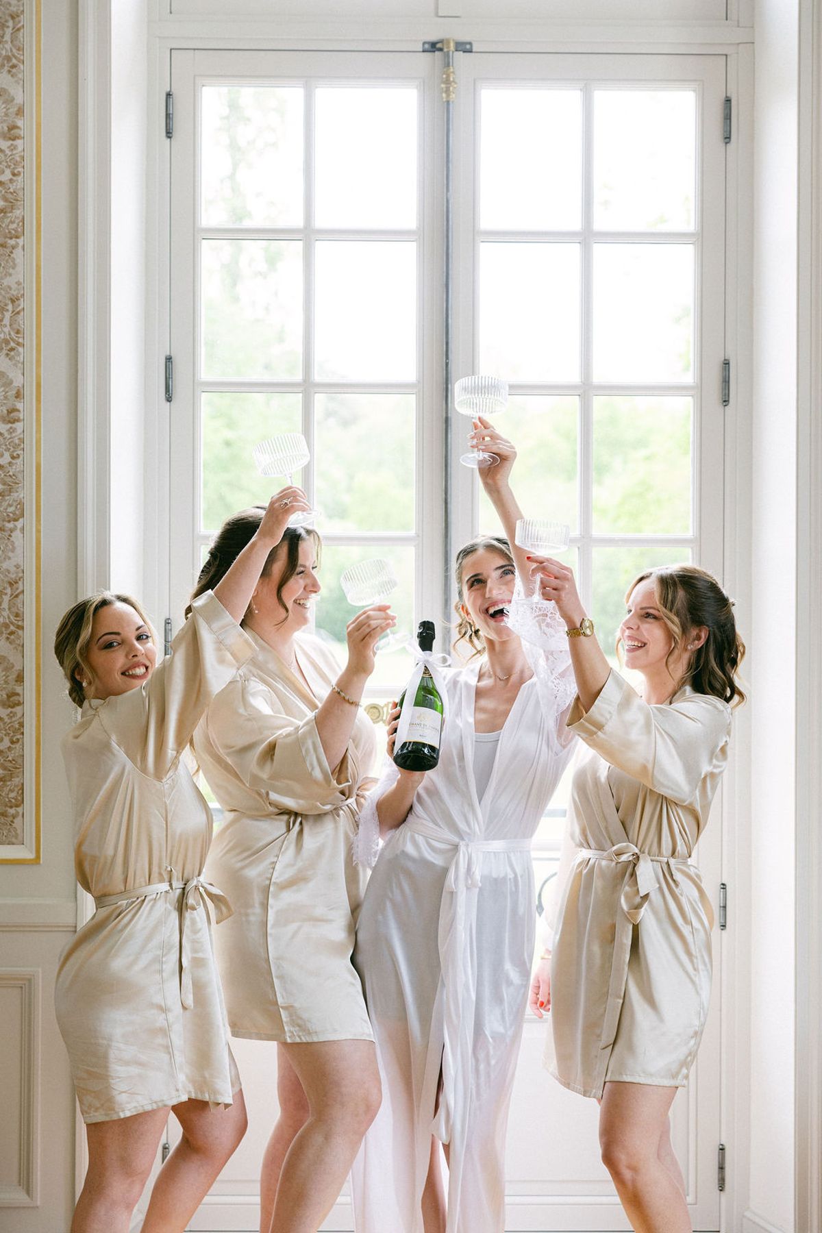 Bridesmaids in robes open champagne in a bright salon at the chateau