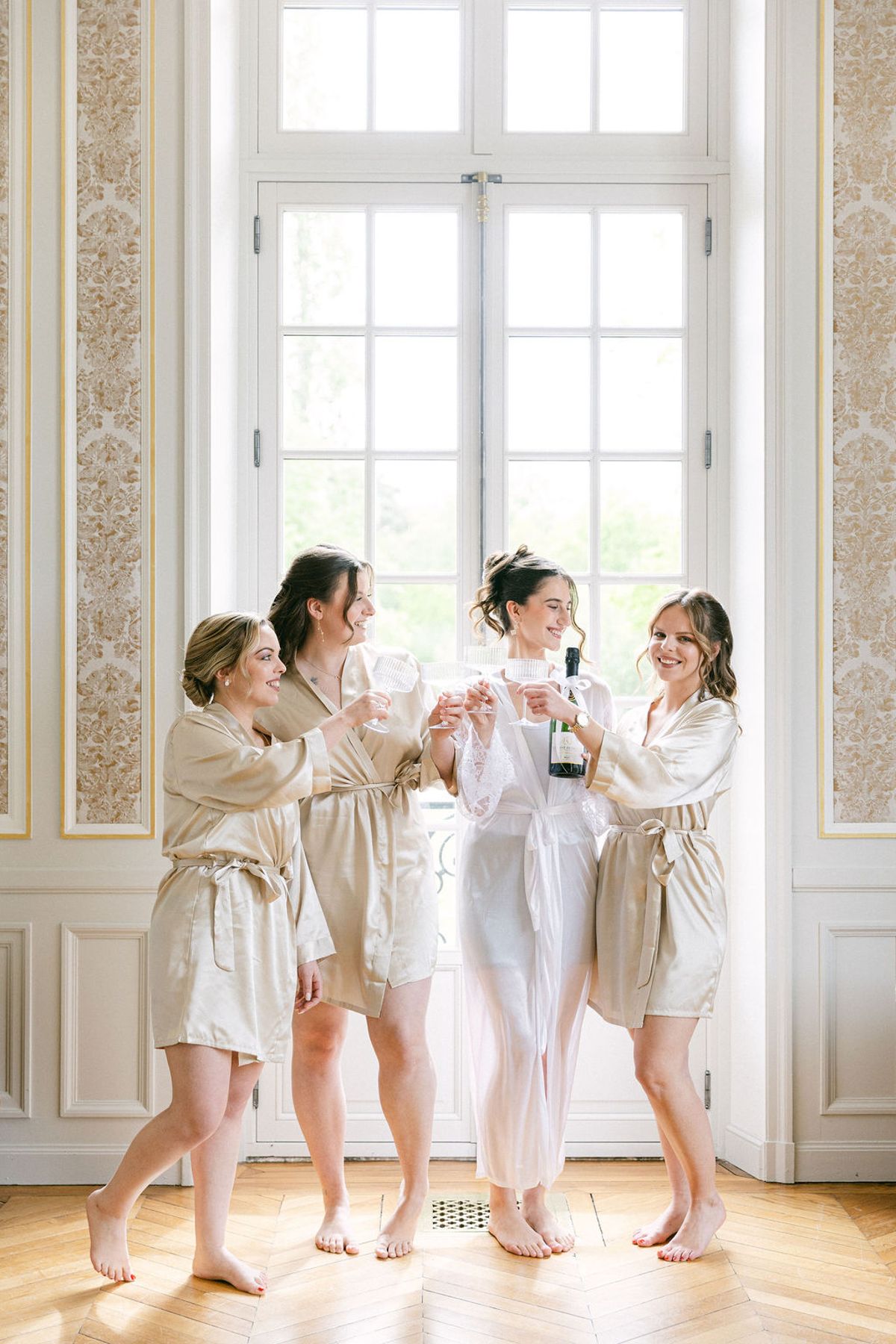 Bridesmaids in robes raise glasses together in a panelled salon at Chateau de Nainville-les-Roches