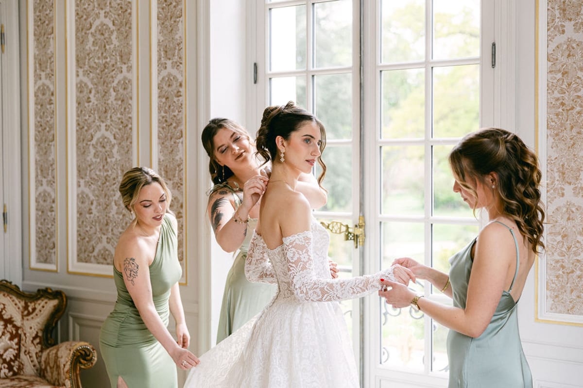 Three bridesmaids in sage green helping the bride into her lace gown at a window