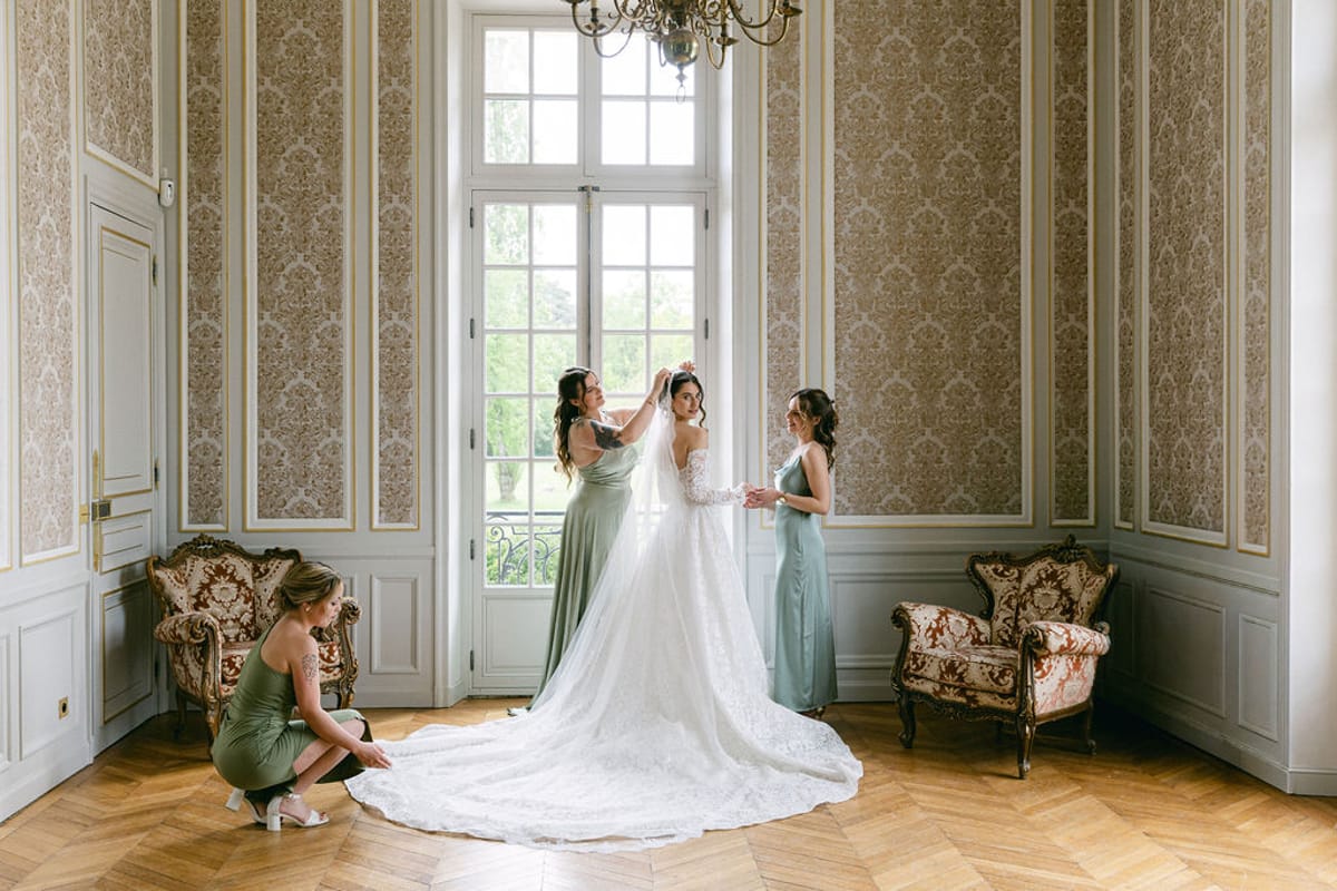 Bride at an open window while one bridesmaid crouches to lay out her train