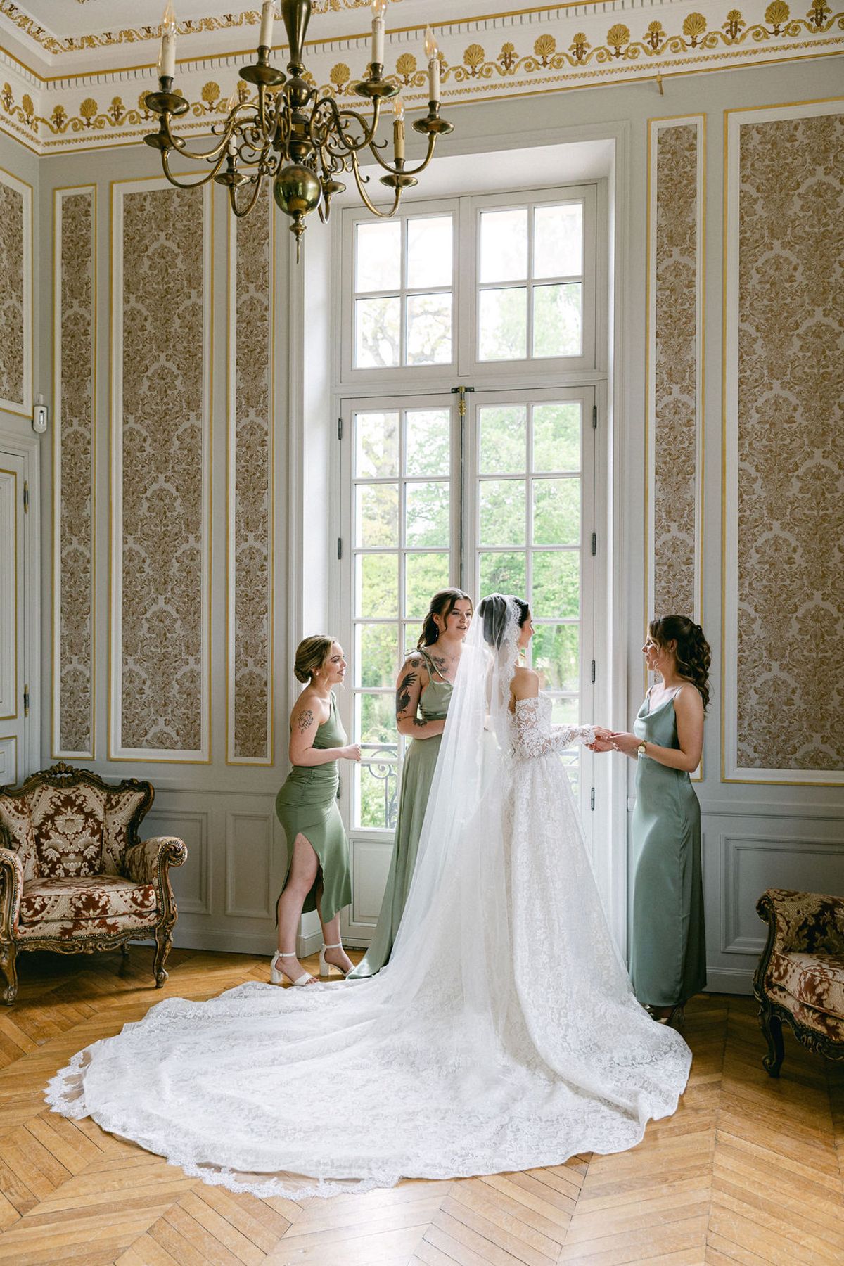 Bride in her veil and train with two bridesmaids beneath a chandelier at a tall window