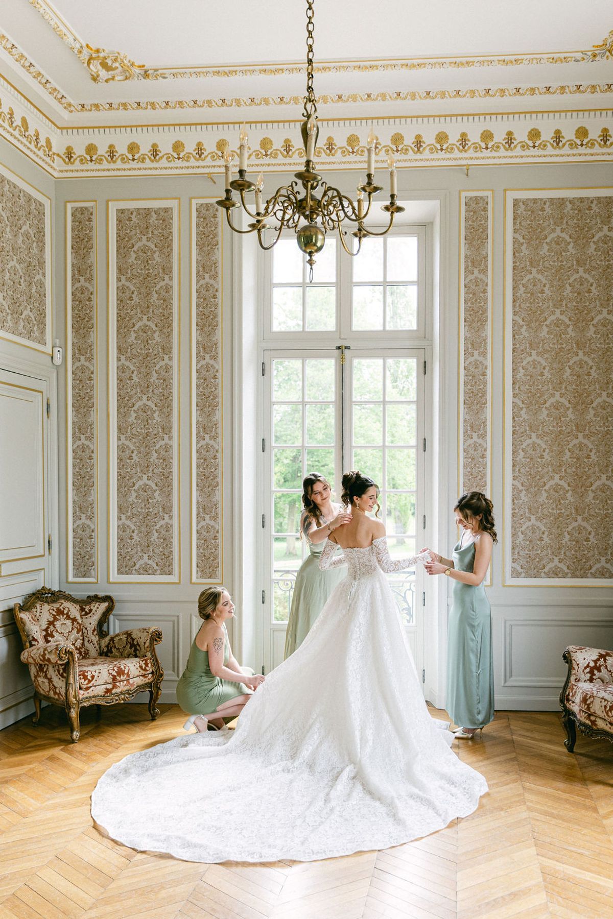 Bride being dressed by her bridesmaids in a wallpapered chateau salon