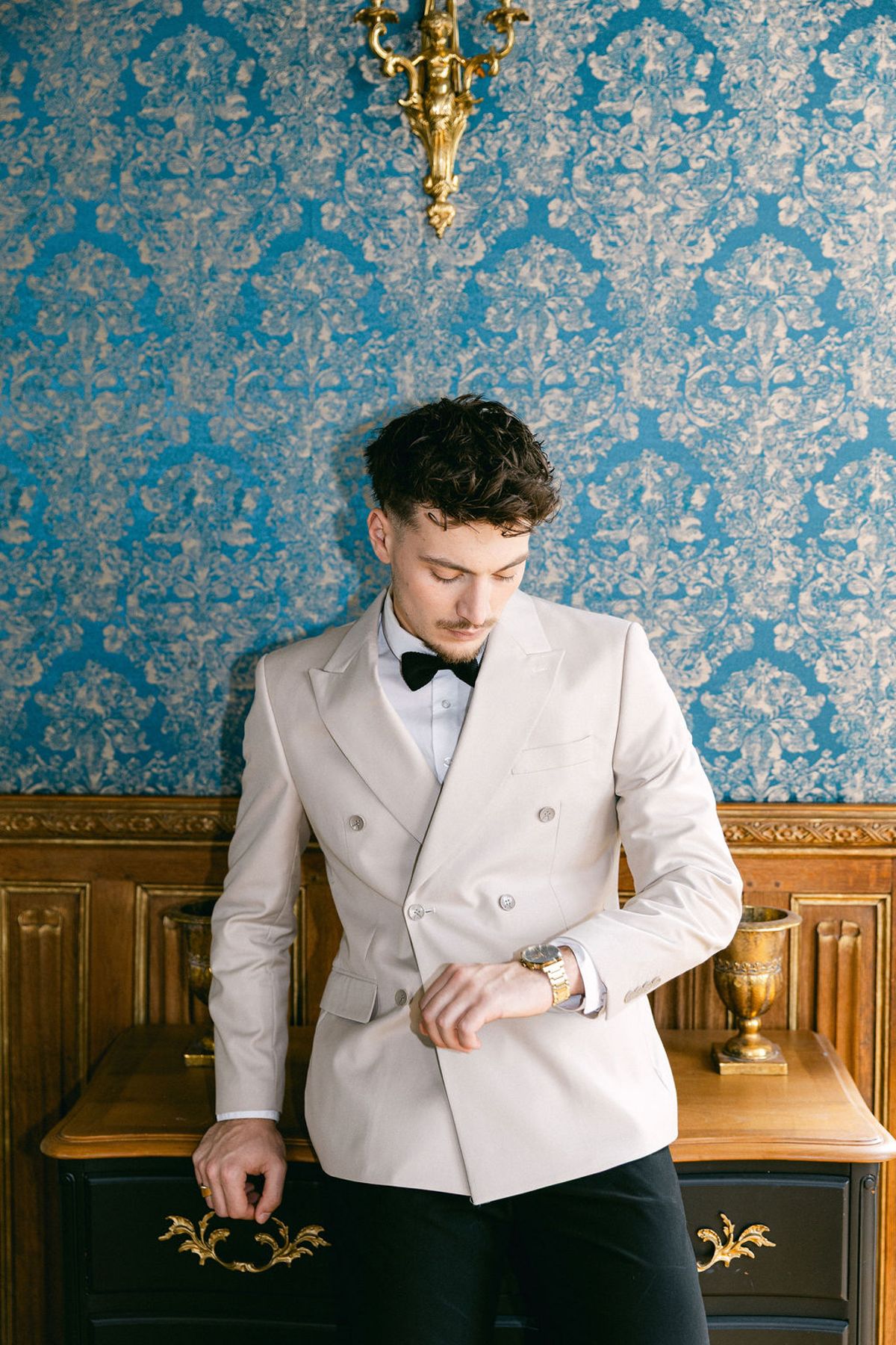 Groom fastens his cream dinner jacket in front of blue damask wallpaper