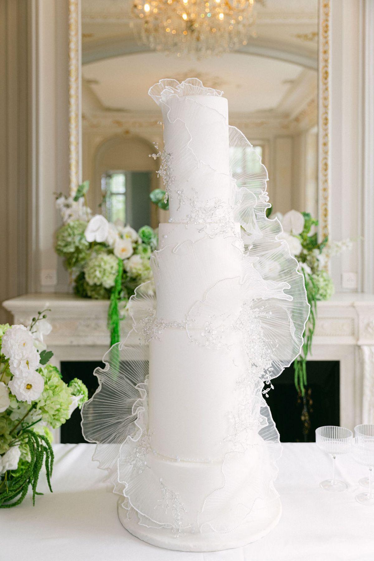 Tall white tiered cake wrapped in pleated wafer-paper fans in a gilded chateau salon