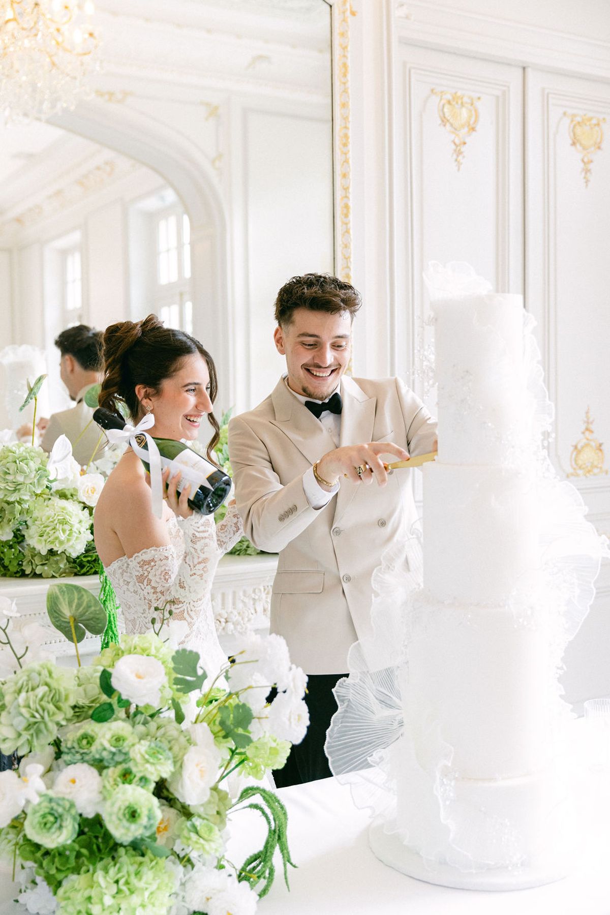 Groom cutting the tiered white wedding cake while the bride holds a bottle of champagne