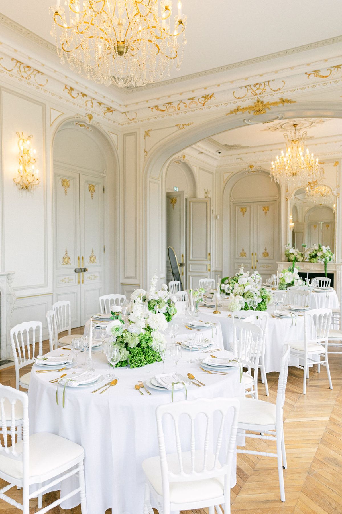 Round reception tables in white linen set beneath a chandelier in a gilded chateau salon