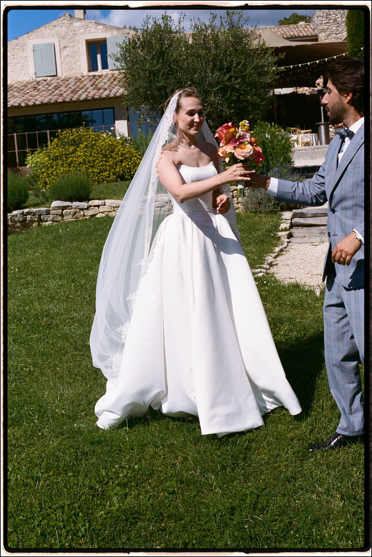 Bride in her wedding gown and veil in a garden, groom nearby