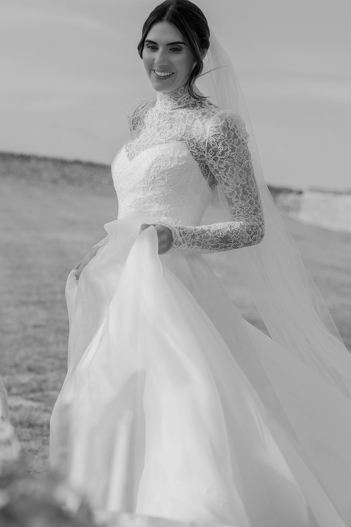 A black and white portrait of a smiling bride in a lace gown and veil, holding up the full skirt of her dress.