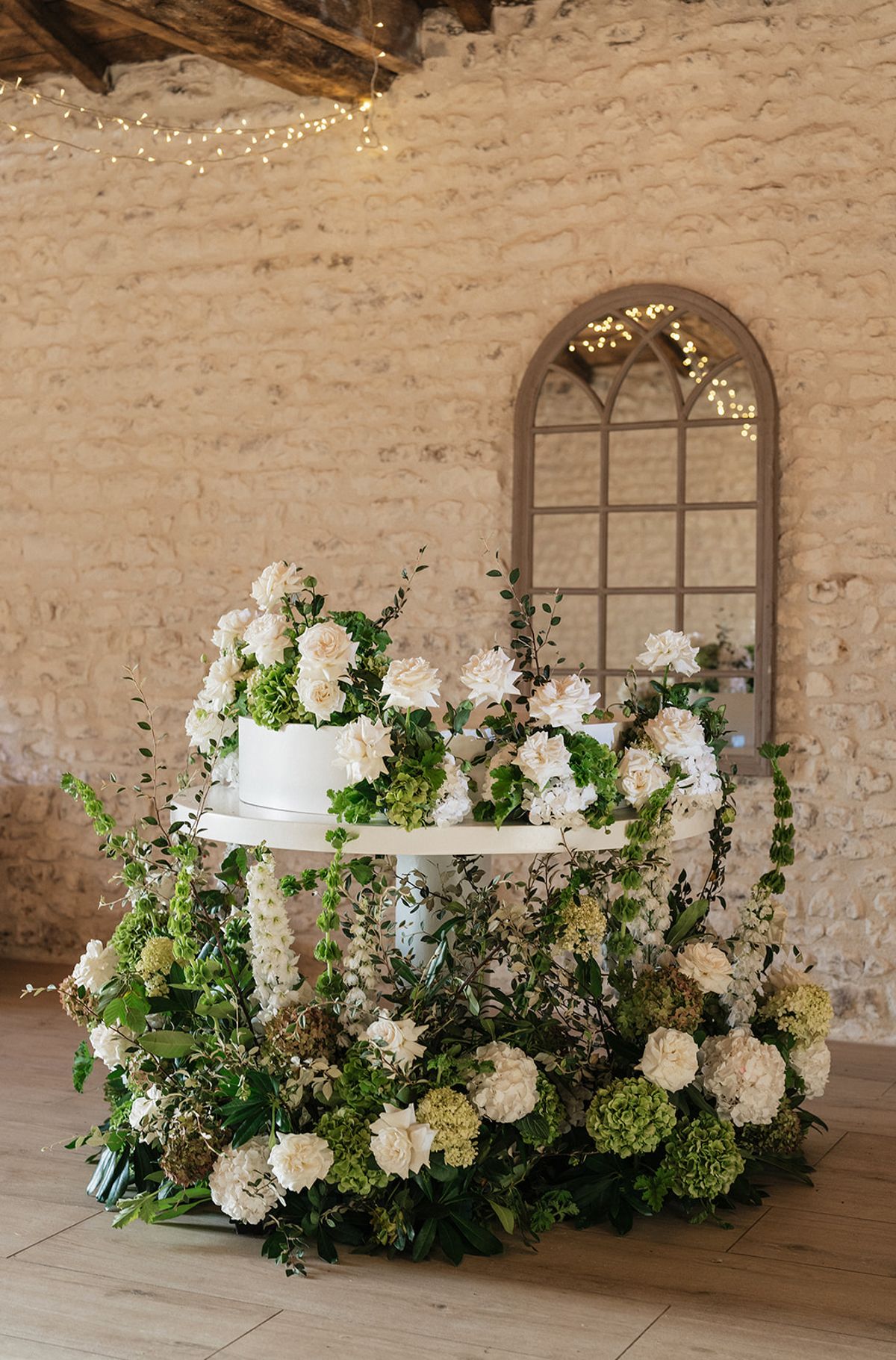 A white cake on a pedestal sits within a lush display of white and green flowers before a stone wall with an arched mirror.