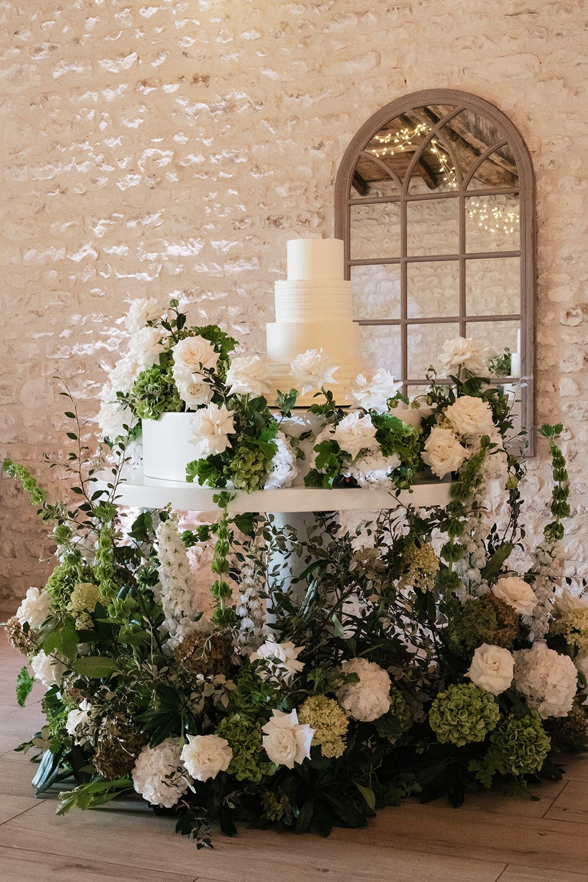 A white tiered cake on a stand is surrounded by green and white flowers in front of an arched mirror and pale brick wall.