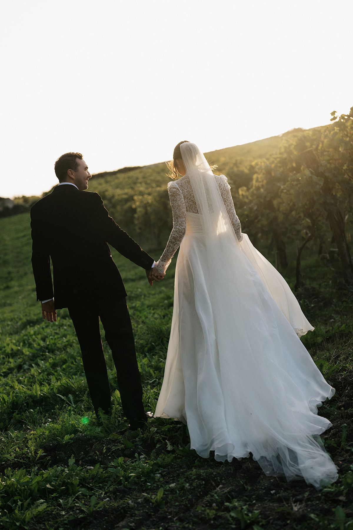 Seen from behind, the couple walk hand in hand across a grassy slope in warm low sunlight, the bride's long veil trailing.