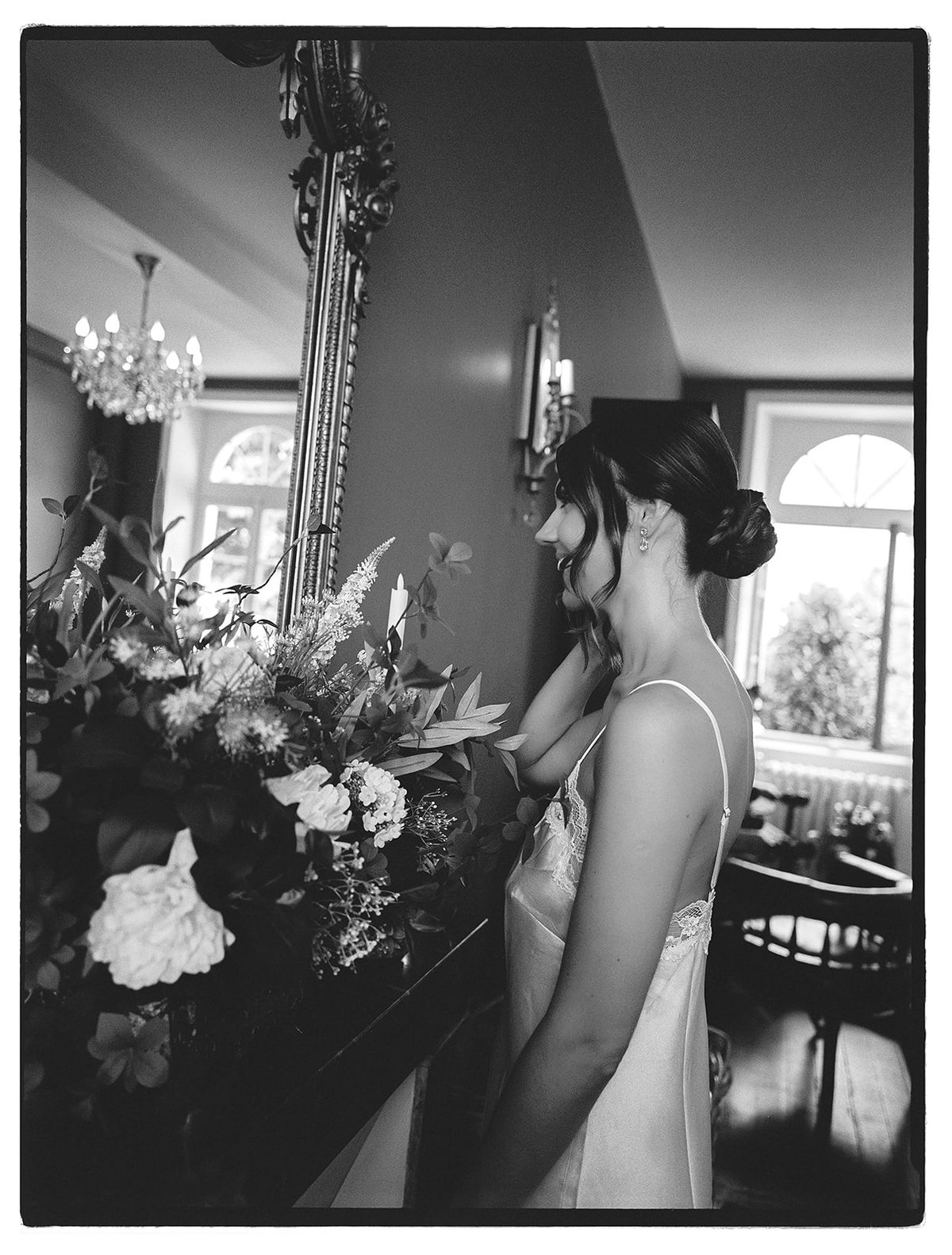 A black and white image of a smiling bride in a slip dress with her hair up, standing beside a large floral arrangement and a mirror.