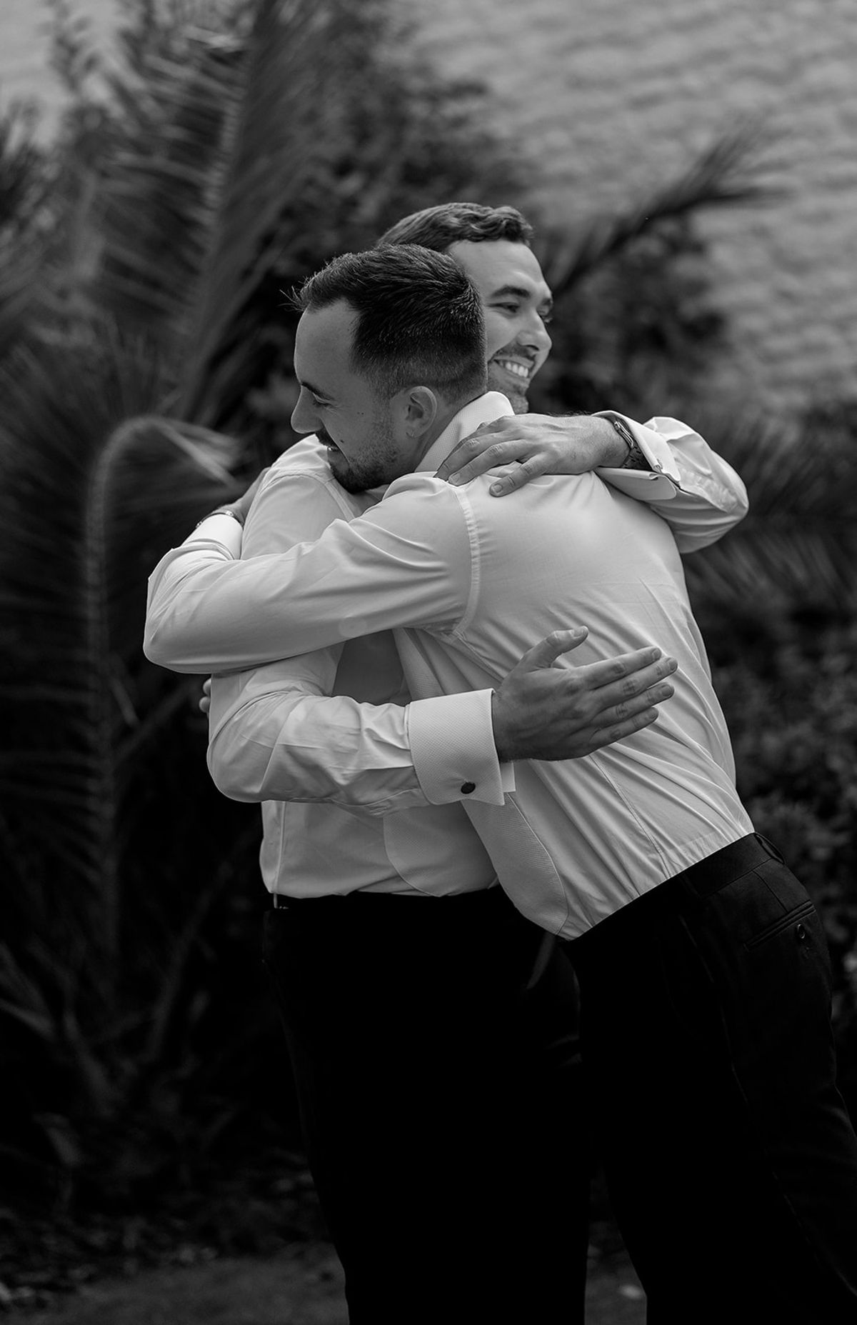 In black and white, two men in white shirts hug closely in front of dark leafy plants.
