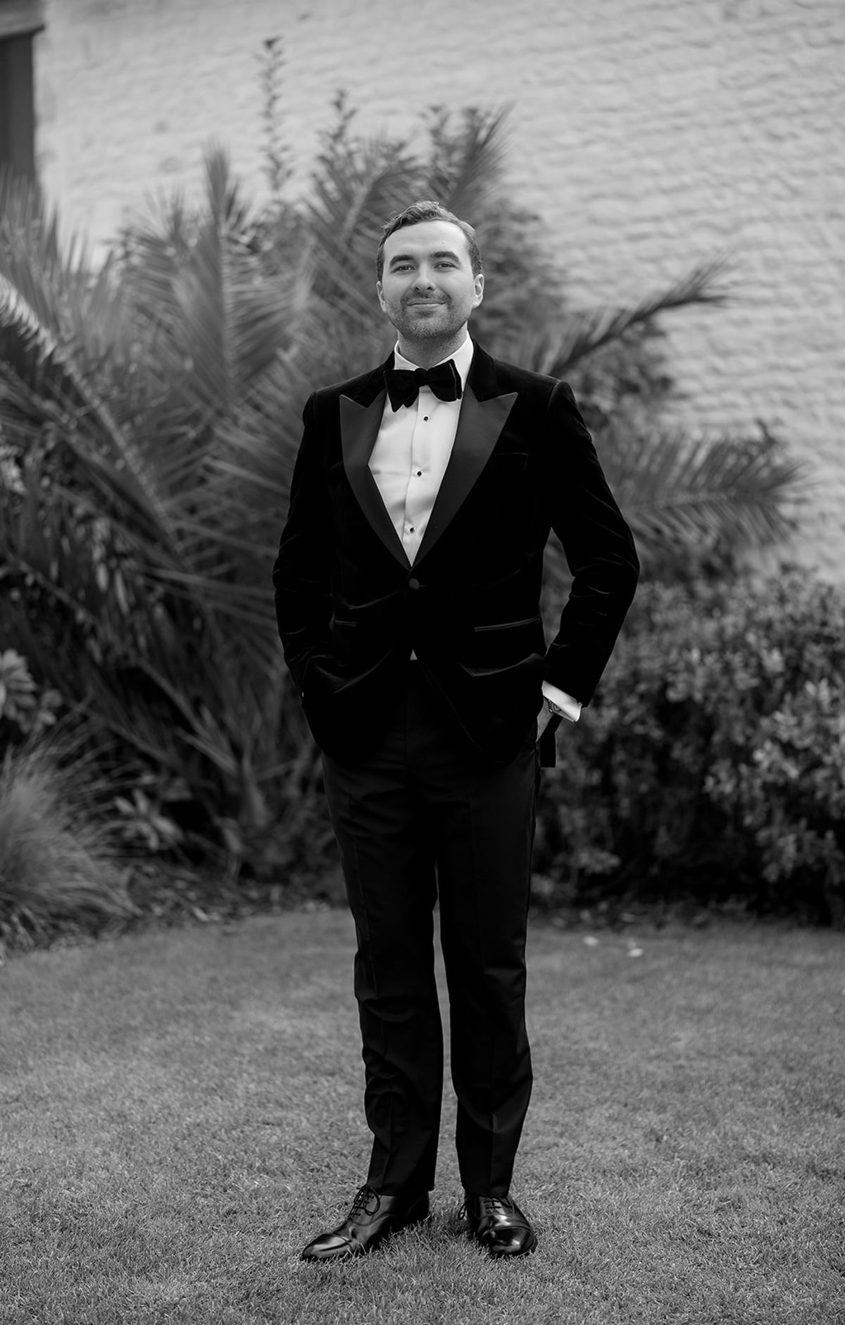 The groom in a dark velvet jacket and bow tie stands smiling with one hand in his pocket in a garden, in black and white.