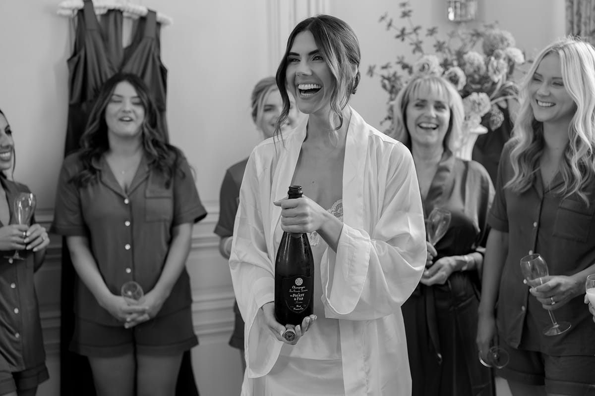 The bride in a white robe laughs while holding a champagne bottle, surrounded by smiling women in pyjamas.