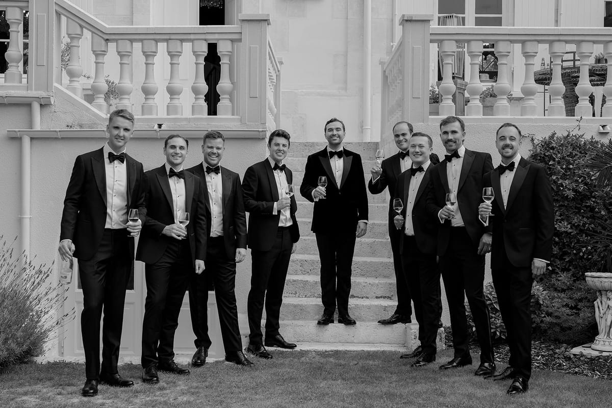 A row of nine men in black tuxedos stand smiling on stone steps in front of a balustrade, in black and white.