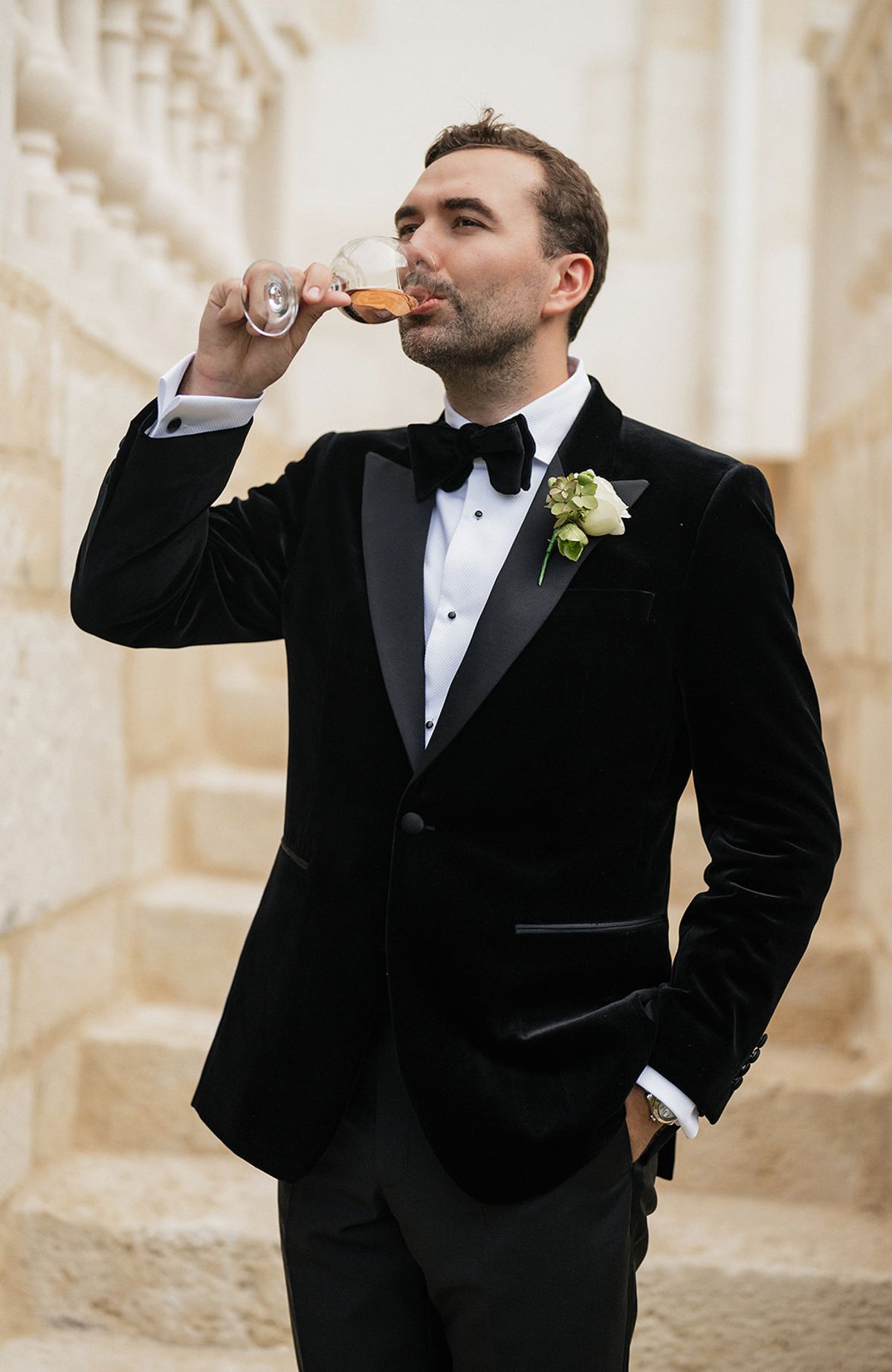 A groom in a black velvet dinner jacket and bow tie sips from a glass beside pale stone steps.