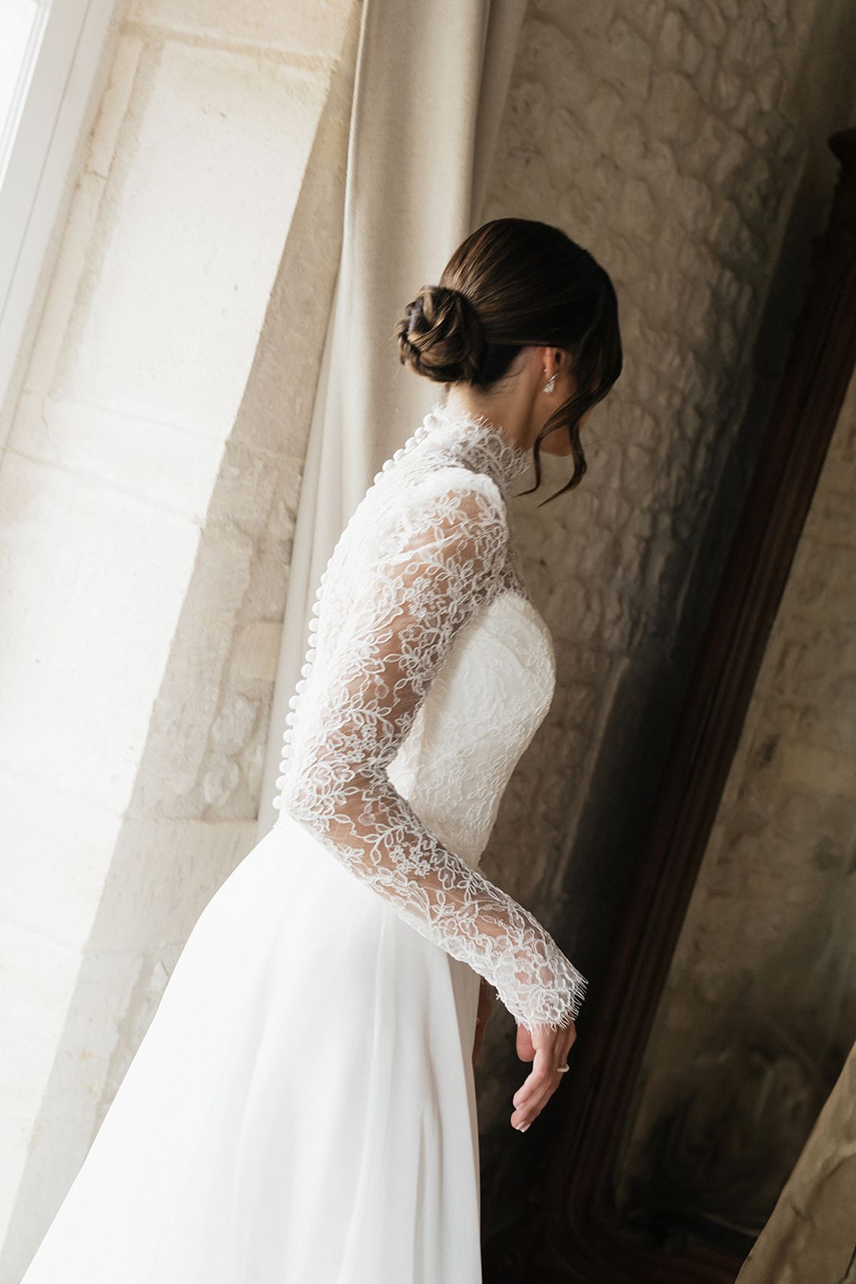 The bride, in a high-necked lace gown with her hair in a bun, stands in profile beside a tall window and stone wall.