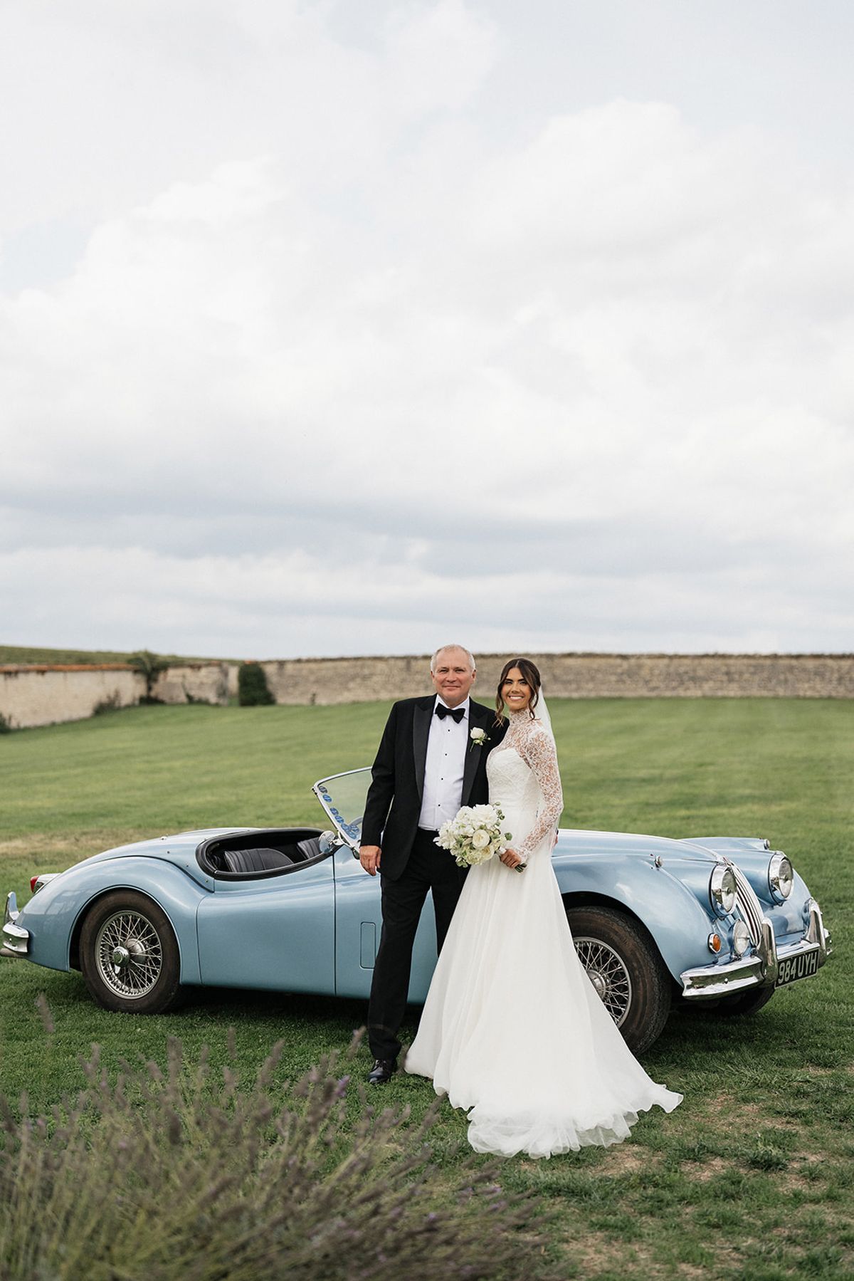 The couple stand together on a wide lawn beside a vintage pale blue convertible under a cloudy sky.