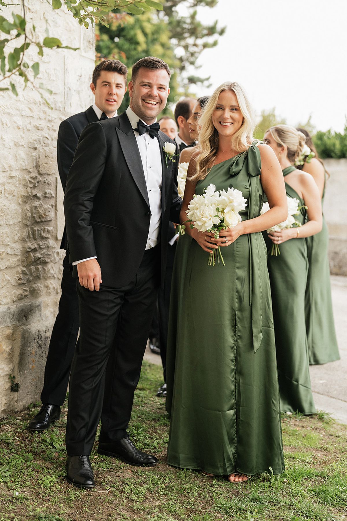 A man in a black tuxedo stands smiling beside a woman in a green satin dress holding white flowers, with others behind them.