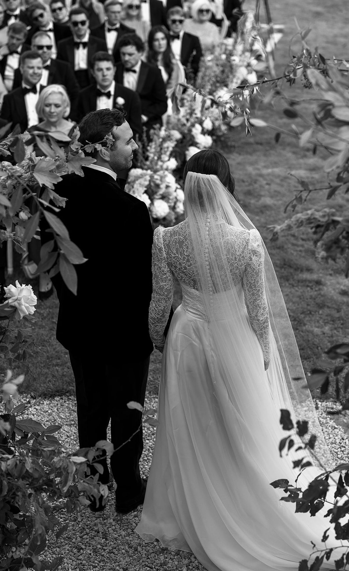 The bride and groom stand with their backs to the camera, facing rows of seated guests, in black and white.