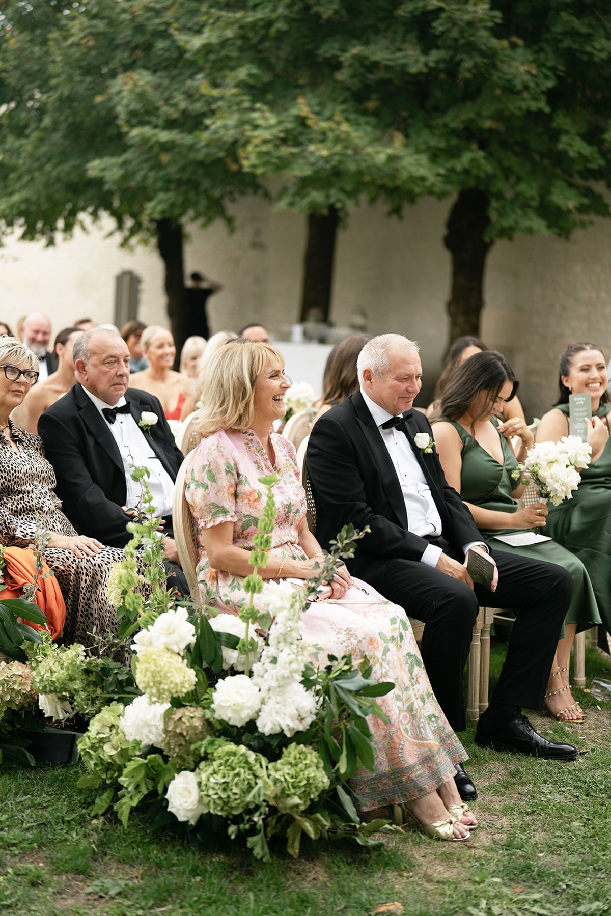 Guests in formal and floral outfits sit in rows under trees next to a large arrangement of white flowers and greenery.