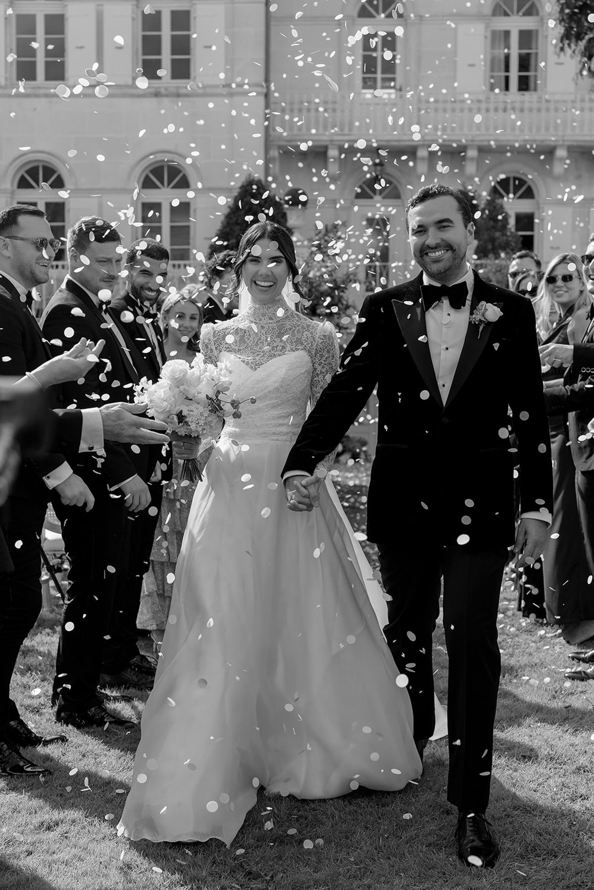 The bride and groom walk hand in hand as guests on either side throw confetti, with a chateau behind them.
