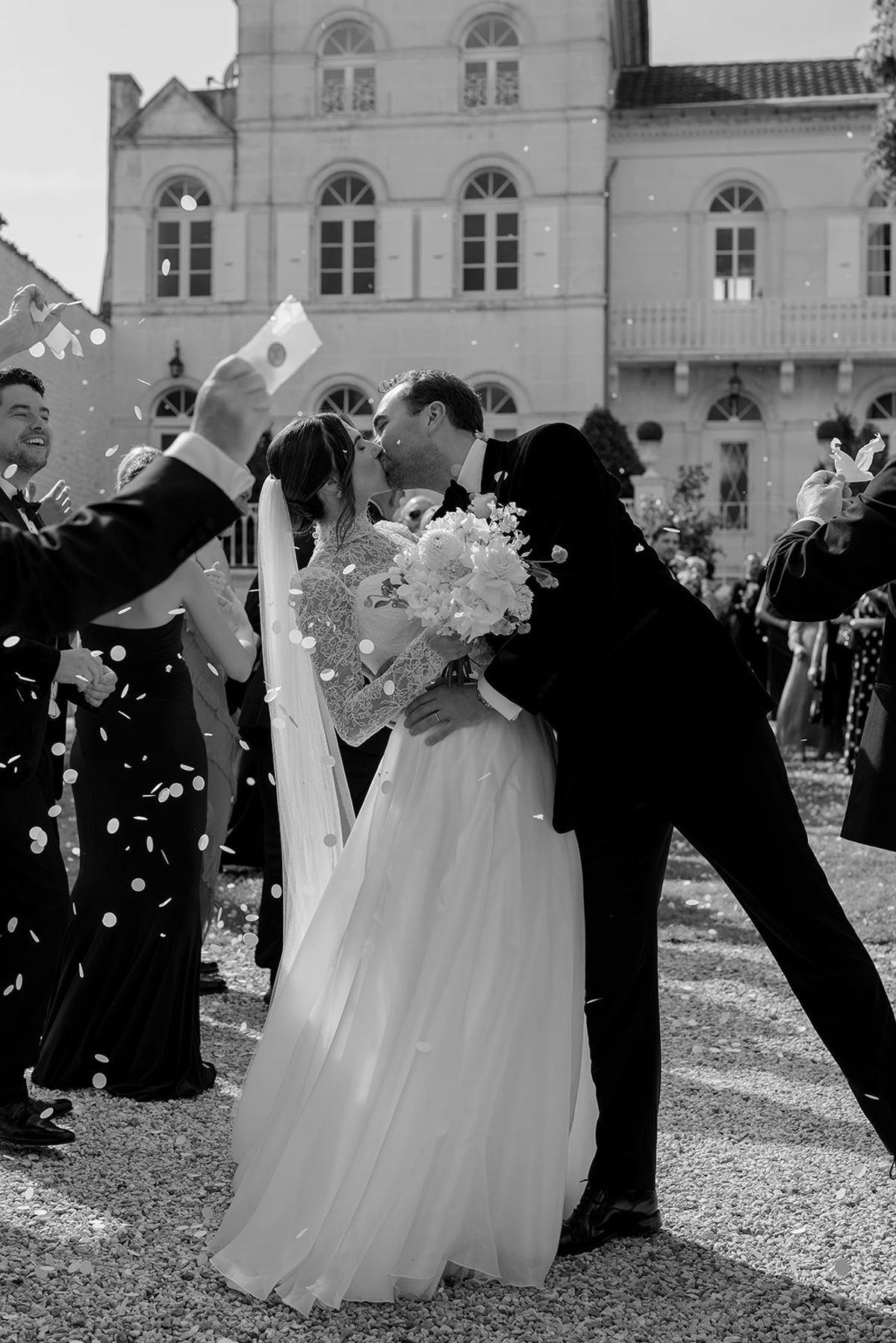 The couple kiss while guests around them throw confetti, with a chateau in the background, in black and white.