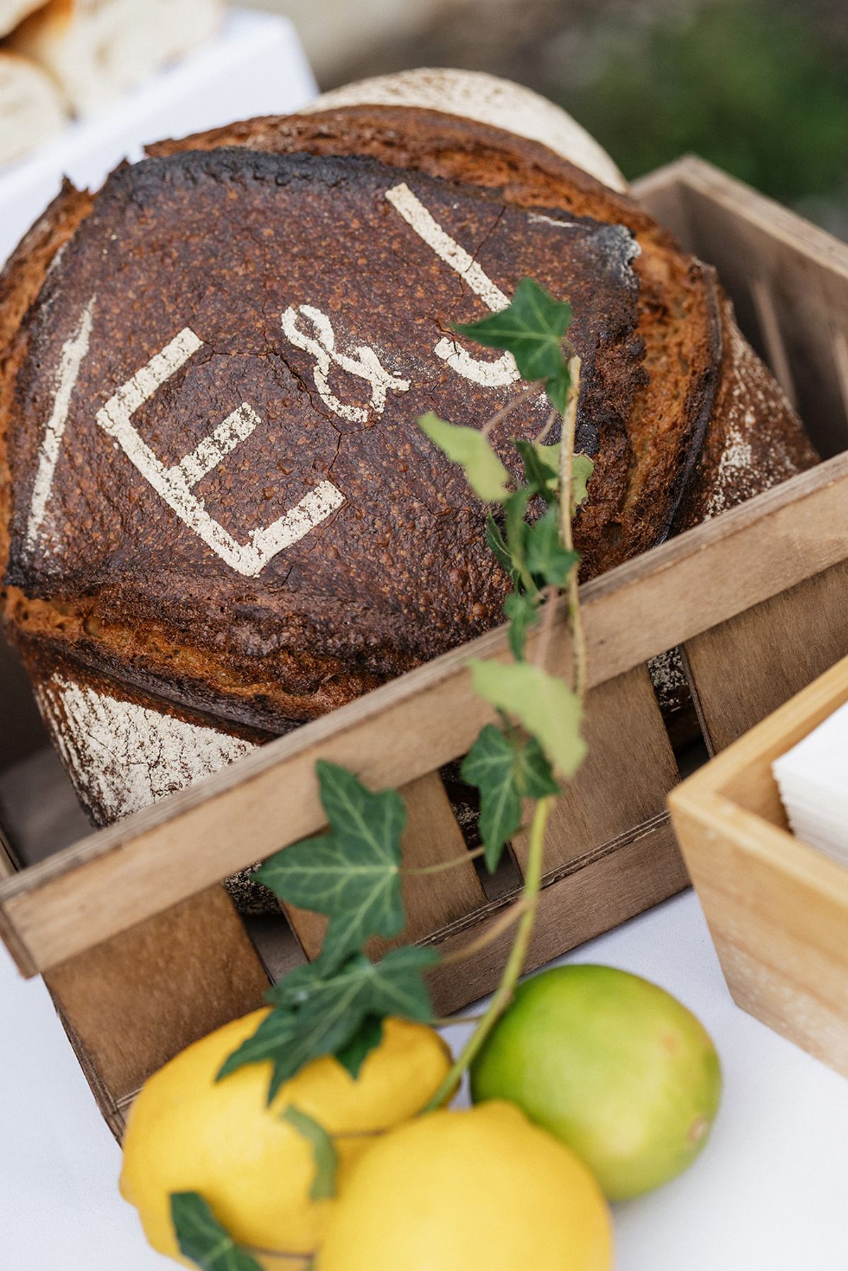 A round loaf scored with two initials sits in a wooden crate with ivy, lemons and limes in the foreground.