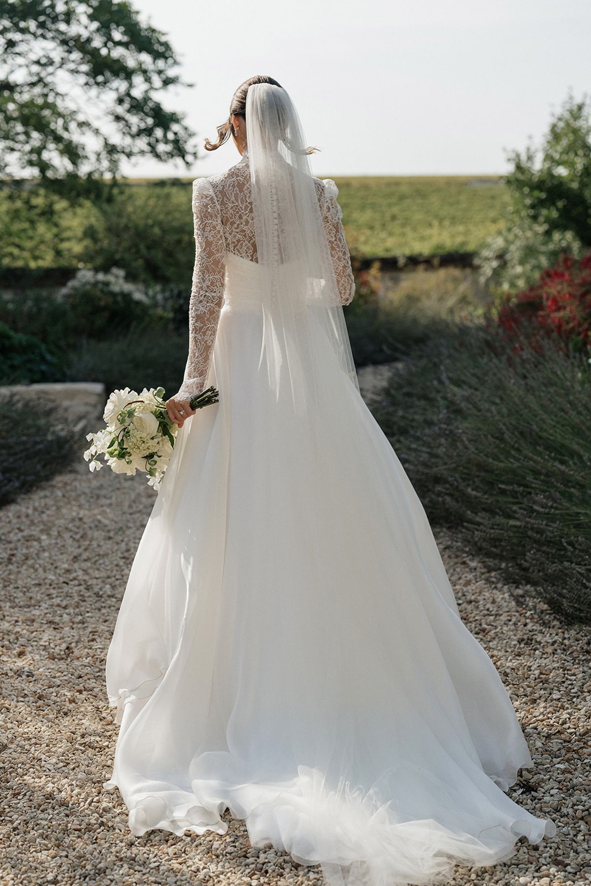 The bride, seen from behind in a long-sleeved gown and veil, walks along a gravel path holding a bouquet beside vines.