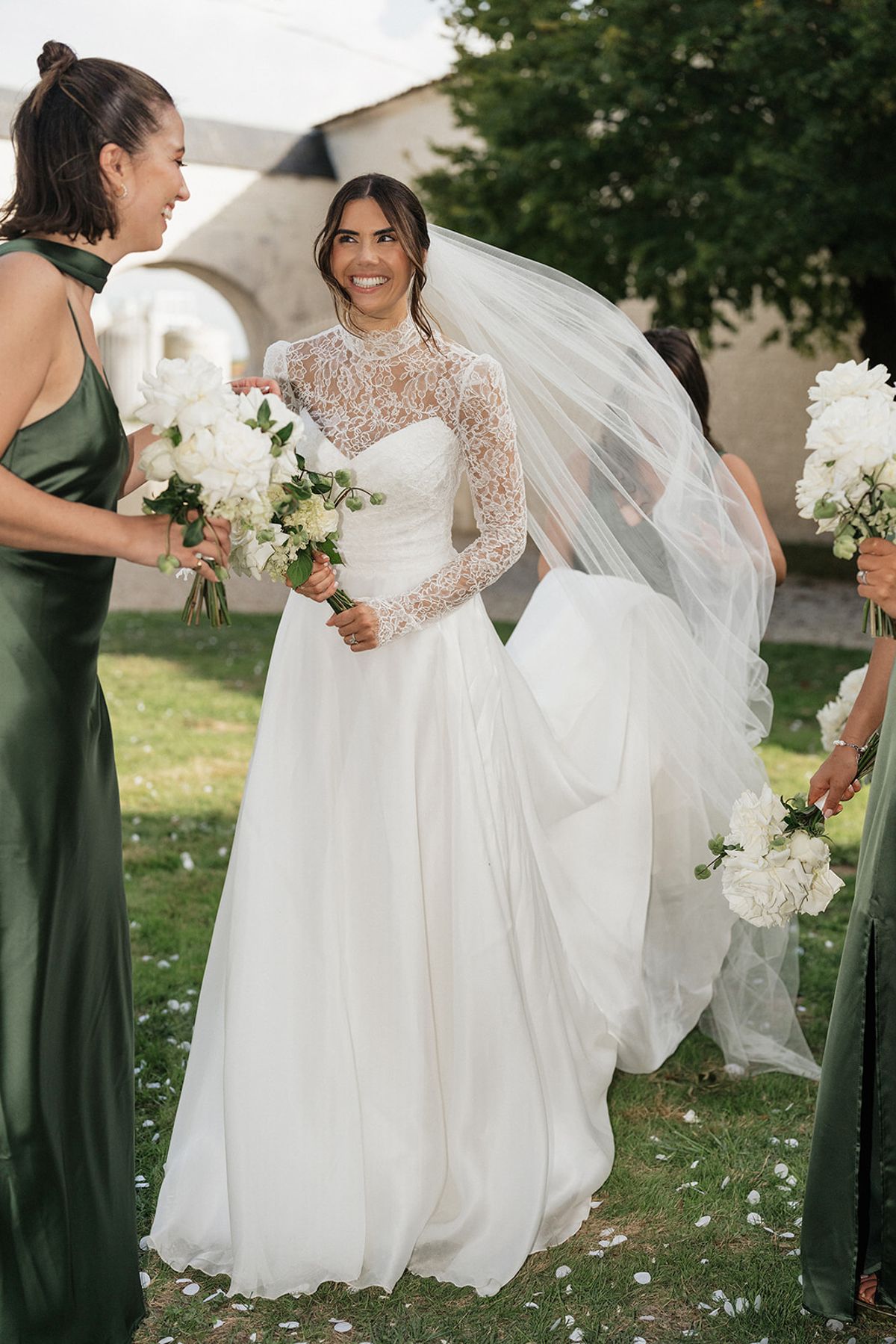The bride in a lace-sleeved gown and veil smiles beside a bridesmaid in a green satin dress, both holding white bouquets on the lawn.