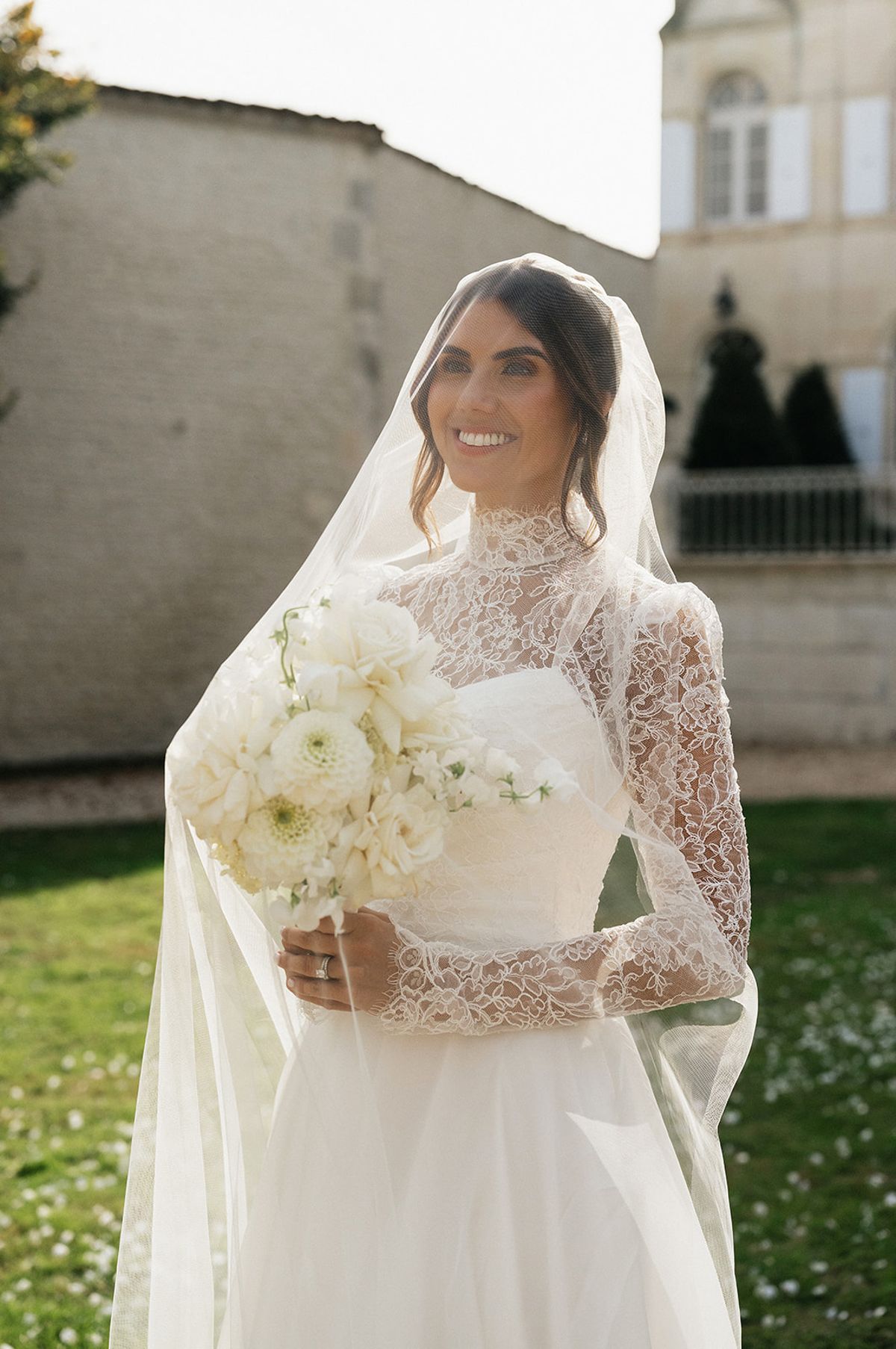 The bride in a high-necked lace gown and long veil smiles while holding a white bouquet on a sunlit lawn.