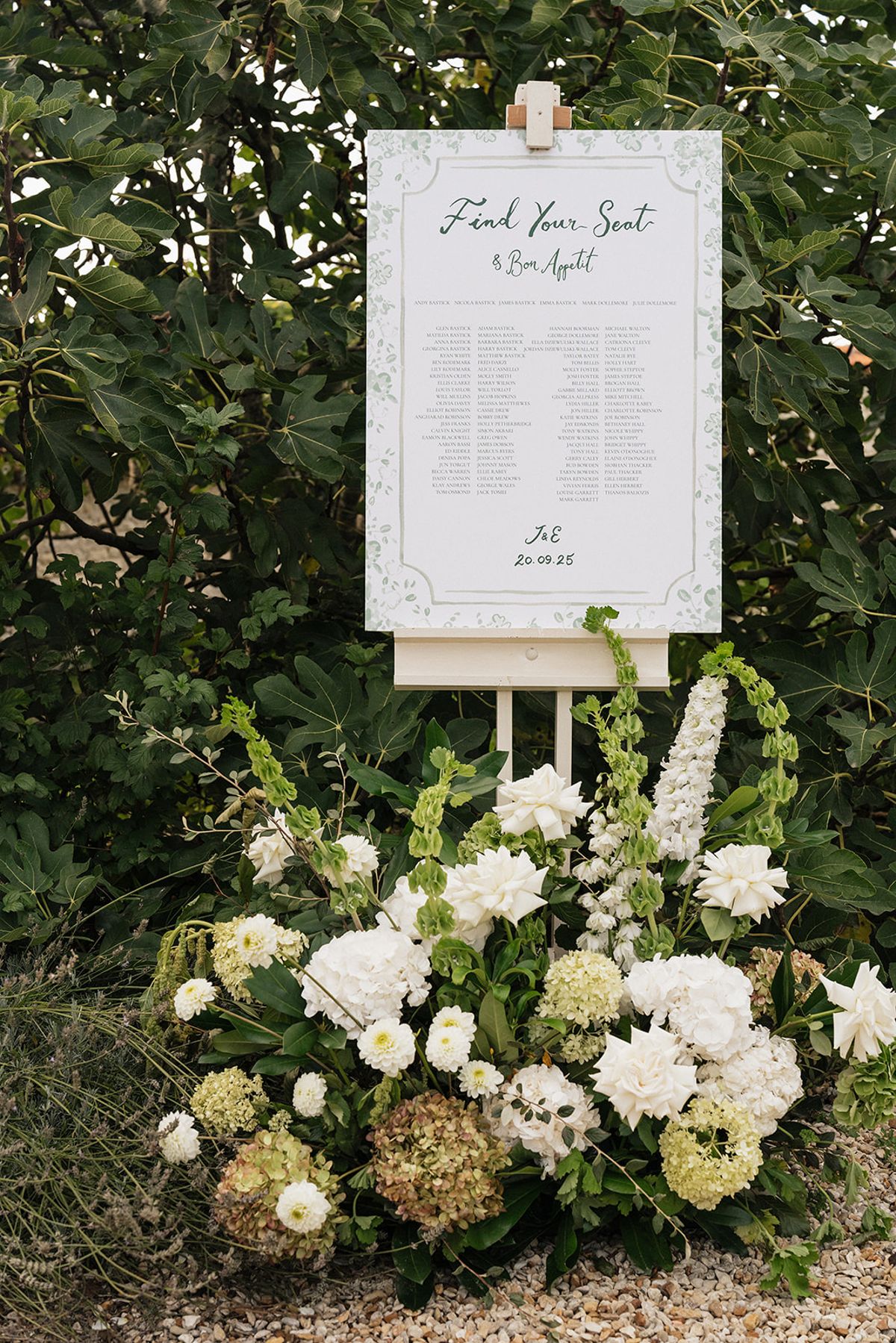A framed seating plan with calligraphy stands on an easel among greenery, above an arrangement of white flowers and hydrangeas.