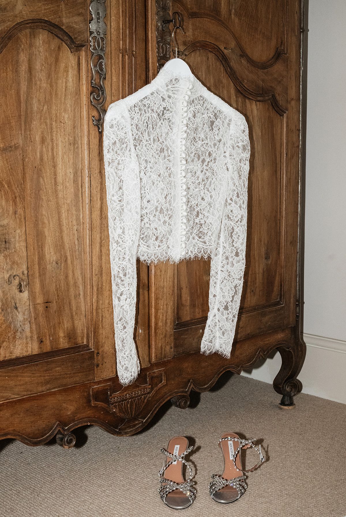 A long-sleeved white lace jacket hangs on a carved wooden wardrobe, with a pair of strappy sandals on the floor below.