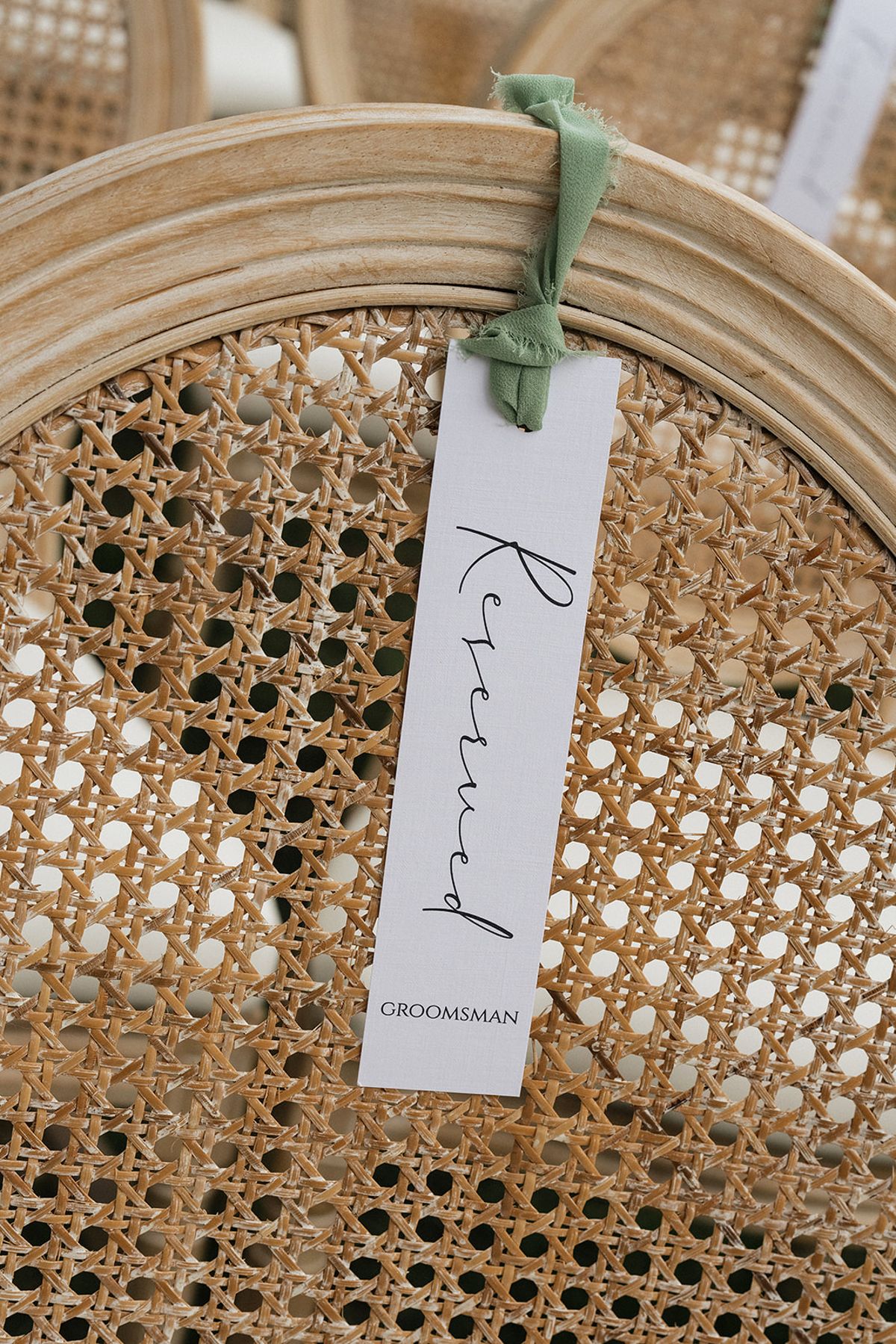 A white reserved card with handwritten-style lettering hangs from a green ribbon on the woven cane back of a wooden chair.