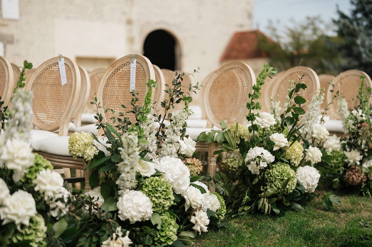 Rows of cane-backed chairs on grass are lined with low arrangements of white hydrangeas and green foliage.