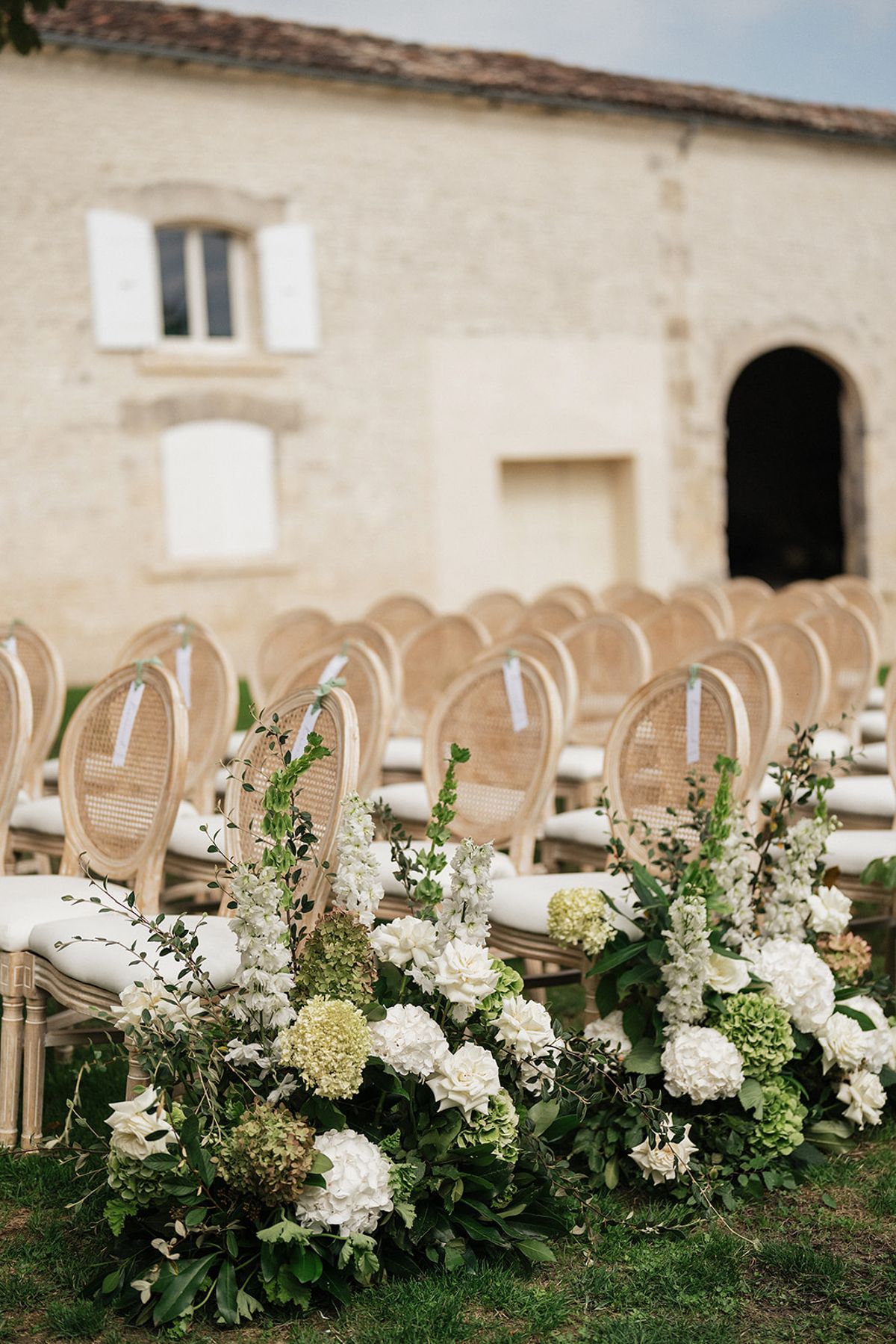 Rows of wooden chairs with rounded backs stand beside tall arrangements of white hydrangeas and flowers in front of a pale stone building.
