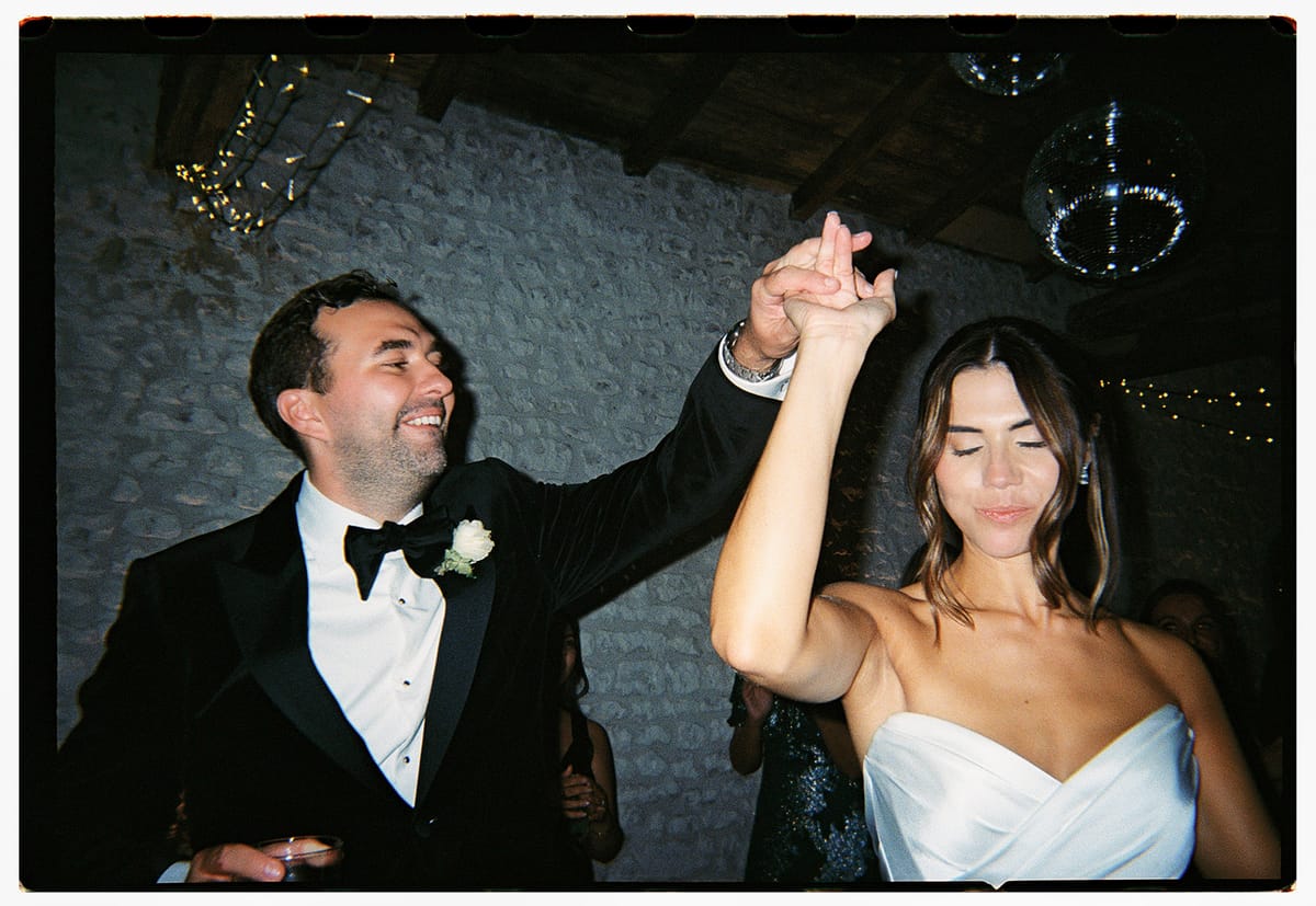 The smiling groom in a dinner jacket and the bride in a strapless dress dance with joined raised hands under string lights.