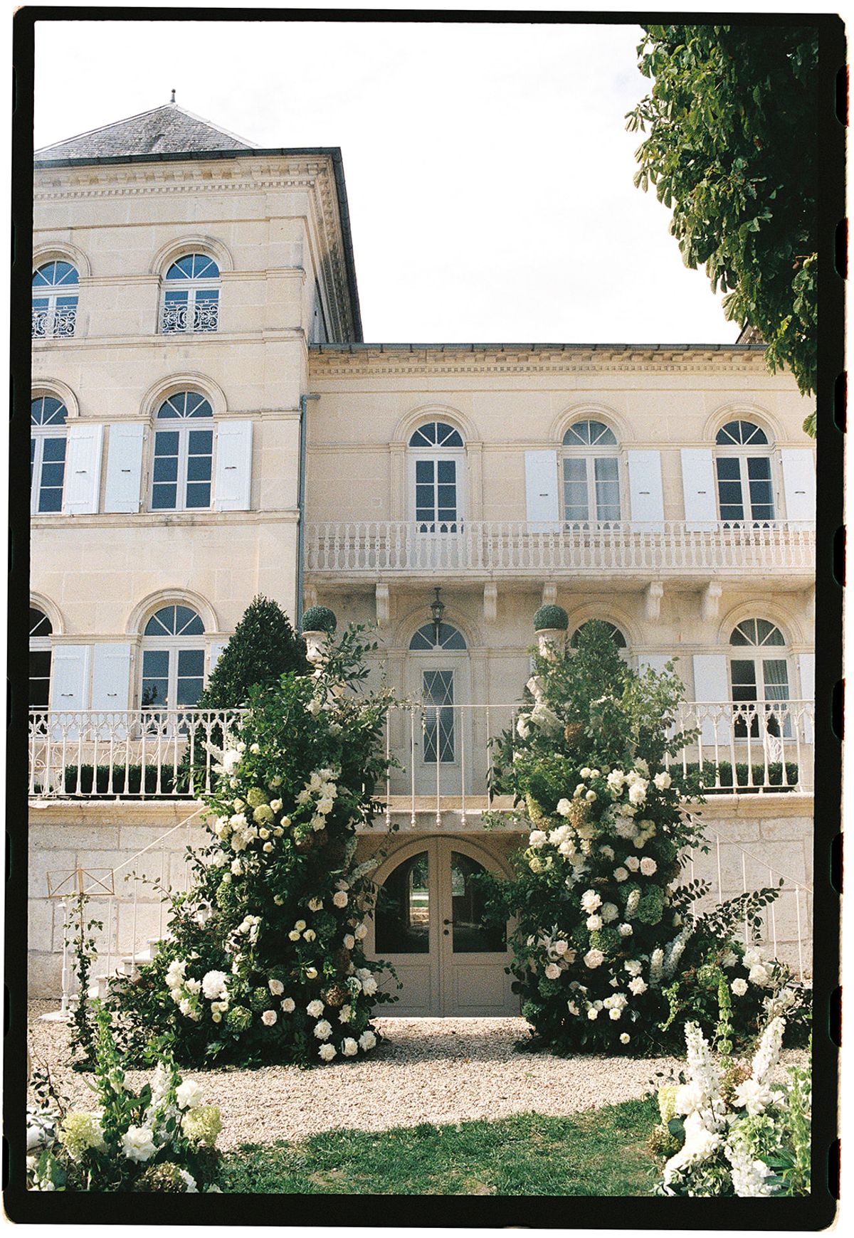 A cream stone chateau with shuttered windows and balconies stands behind two tall white floral arrangements on the lawn.
