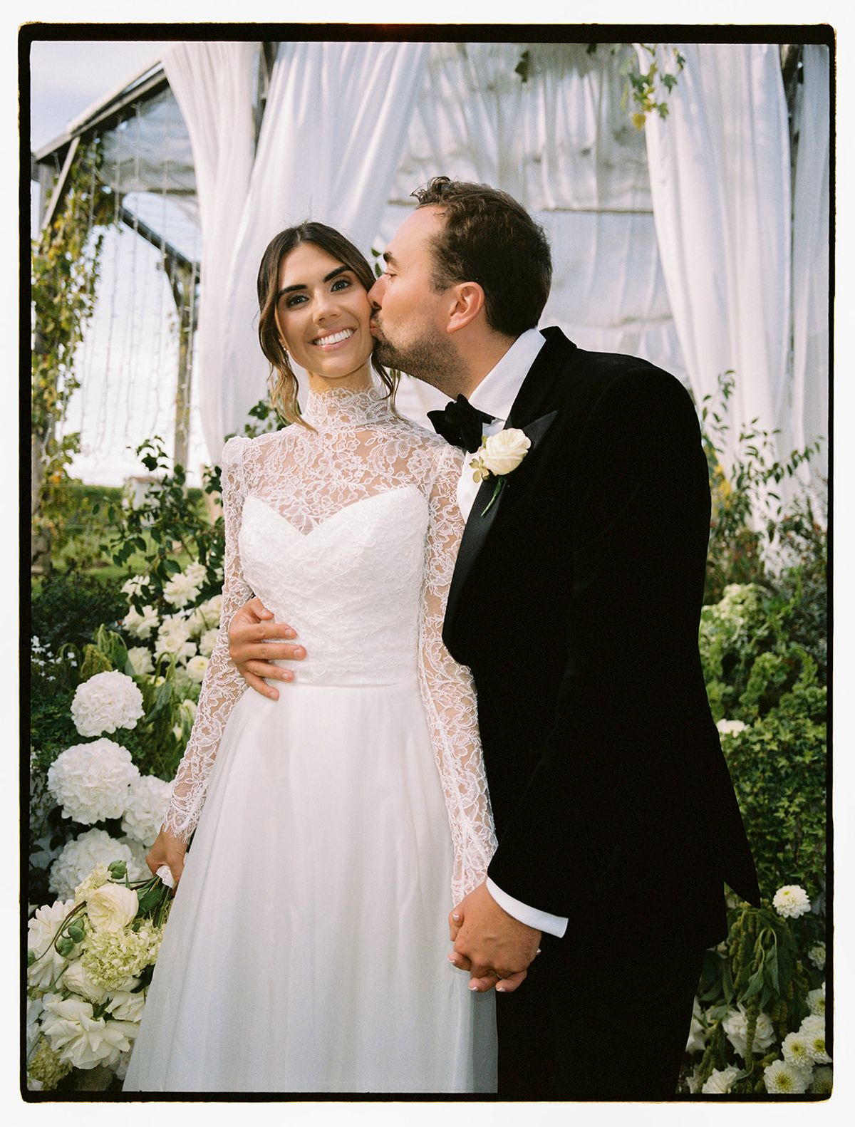 The groom kisses the bride on the cheek as they embrace, she in a lace high-necked gown, surrounded by white flowers and drapes.