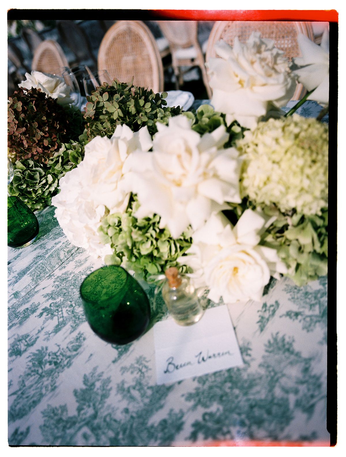 A centrepiece of white roses and hydrangeas sits on a green and white patterned tablecloth beside a green glass and a handwritten place card.
