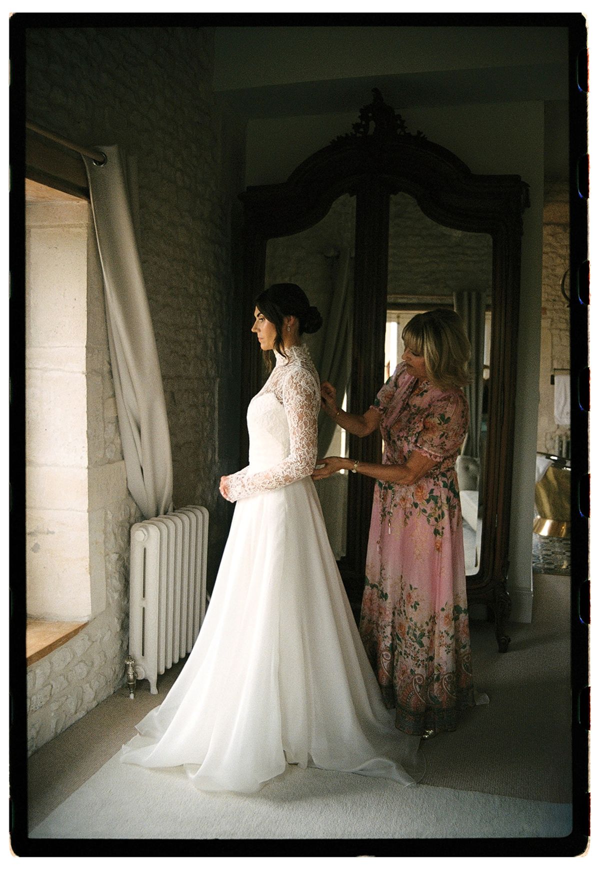 A woman in a floral dress fastens the back of the bride's gown in a stone room with a tall wardrobe mirror.