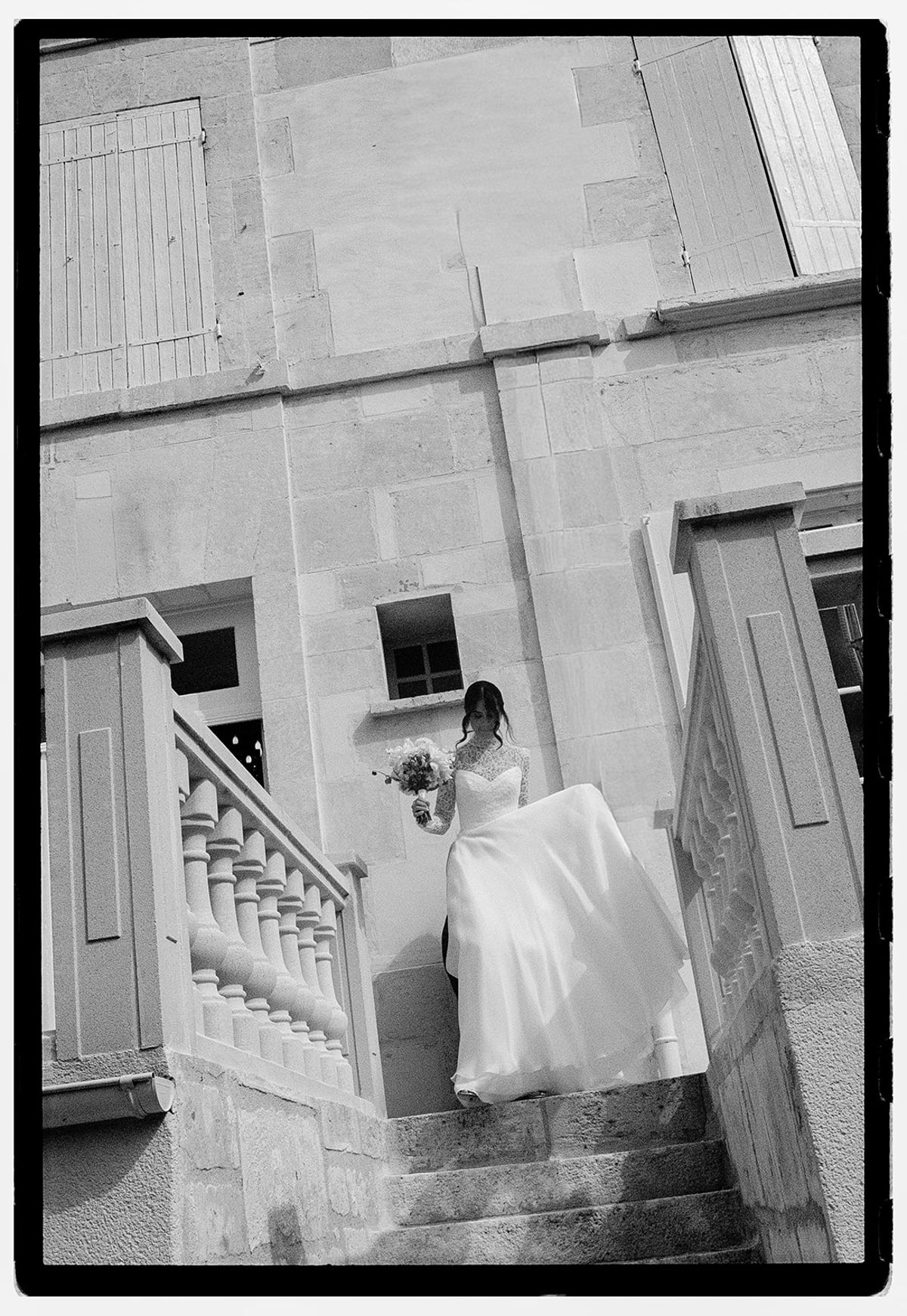 In black and white, the bride in a long gown holds a bouquet as she walks down a stone staircase with balustrades.
