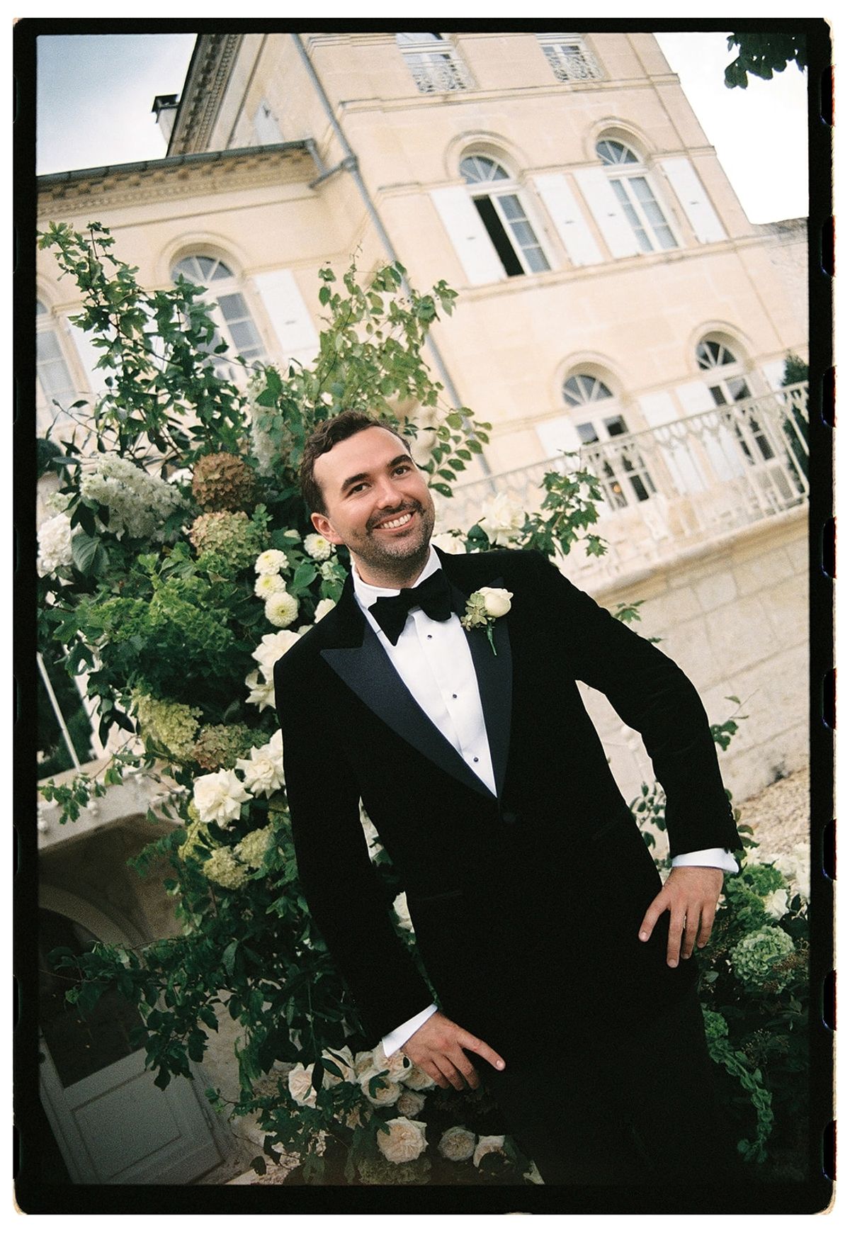 A smiling groom in a black tuxedo leans against a lush green and white floral arrangement in front of a pale chateau.