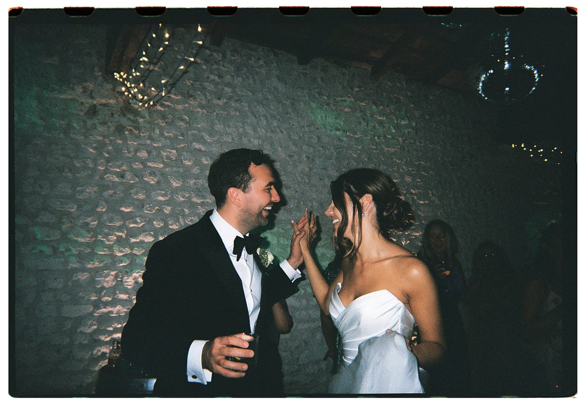 The couple laugh and dance together in a dimly lit stone-walled room with string lights, framed by a dark film-style border.